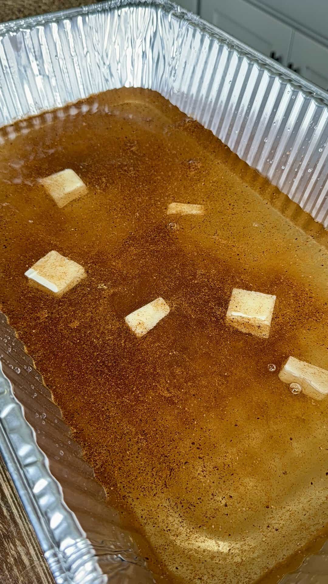 A large aluminum tray filled with seasoned liquid and several small cubes of butter floating on the surface. The liquid appears to have a reddish-brown tint, suggesting spices or broth.