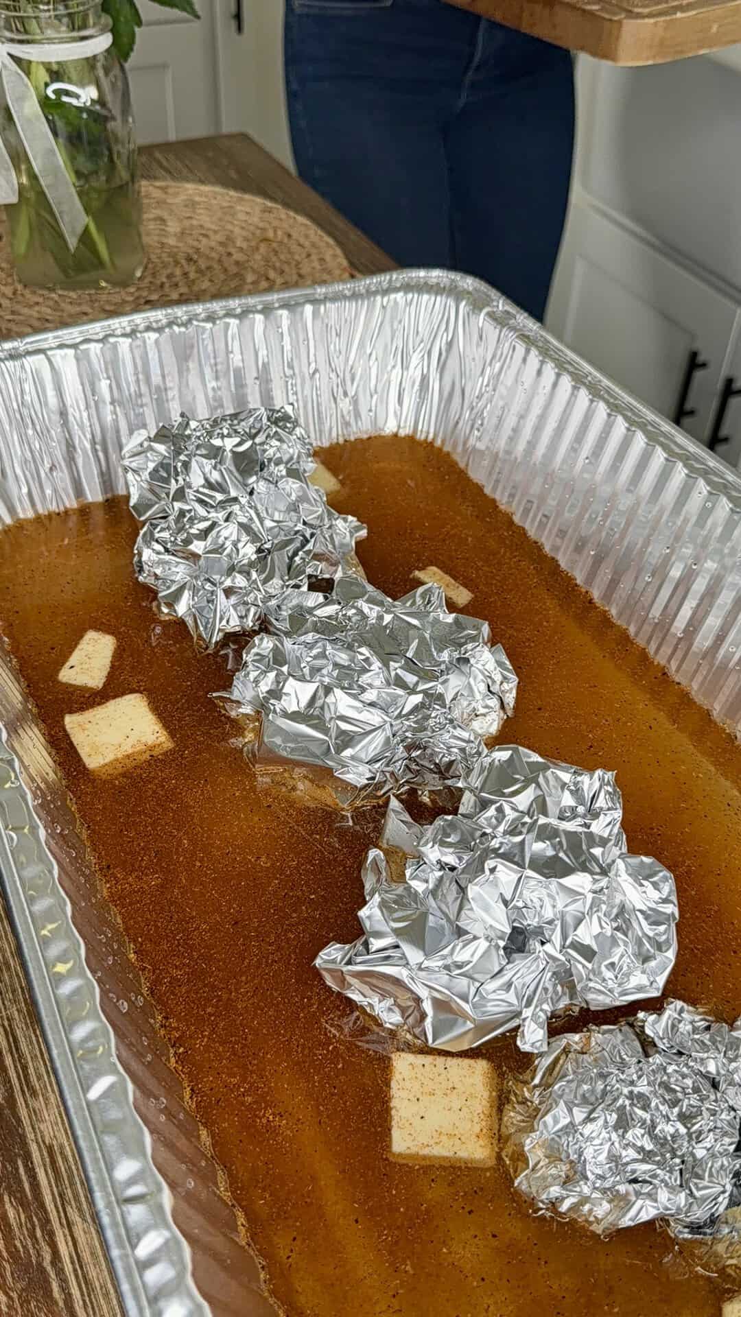 Four foil-wrapped bundles and butter slices in a large aluminum tray filled with seasoned liquid, sitting on a kitchen counter with a person holding a cutting board in the background.