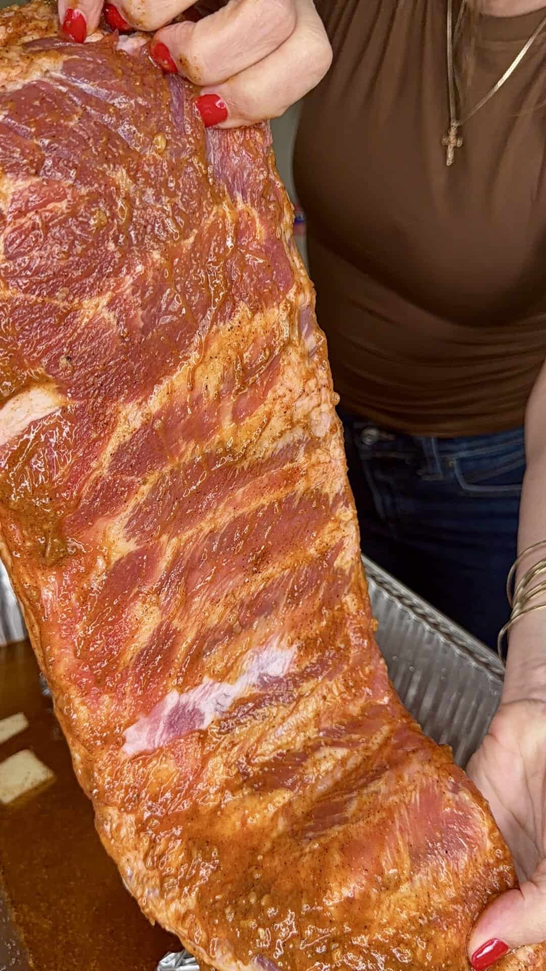 A person with red nail polish holds up a large, seasoned rack of raw ribs over a metal tray, preparing it for cooking.