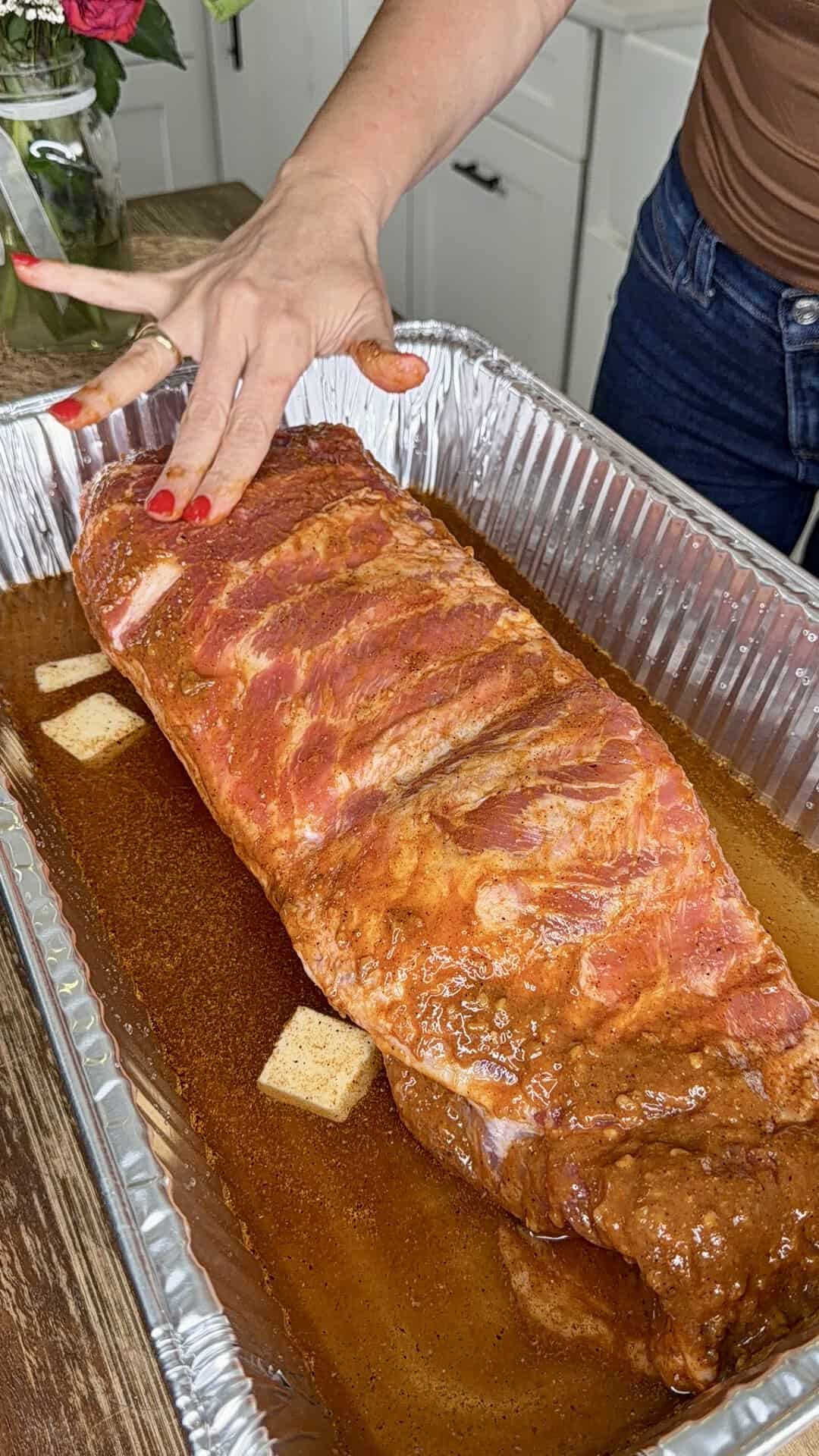 A person with red nail polish is seasoning a large raw rack of ribs in a foil tray, with brown seasoning cubes and marinade at the bottom. The scene is set in a kitchen.