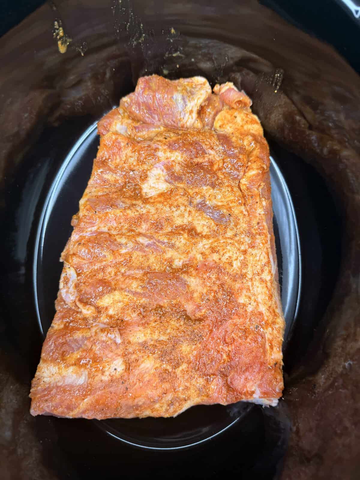 A slab of raw, seasoned pork ribs sits inside a black slow cooker, ready to be cooked. The ribs are coated with a reddish spice rub.