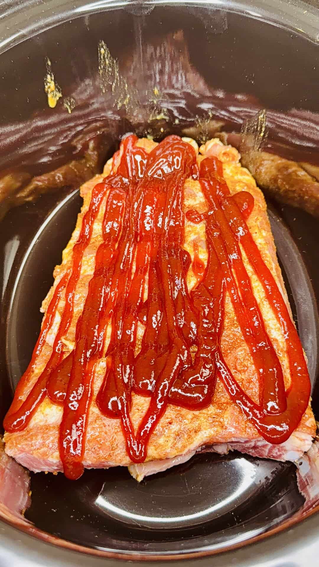 A raw meatloaf topped with ketchup sits inside a slow cooker, ready to be cooked. The ketchup is spread in thick lines across the top of the meat mixture.