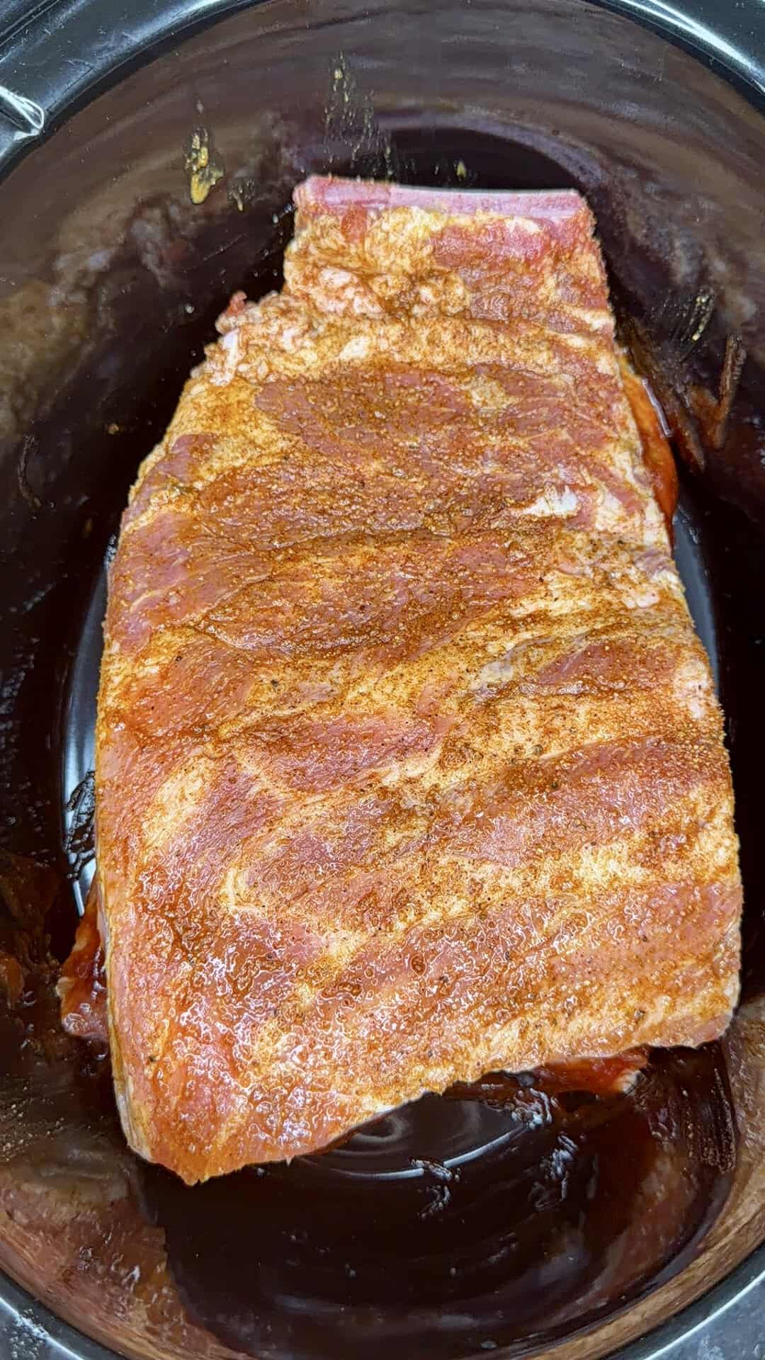 A slab of seasoned raw pork ribs sits in a slow cooker, ready to be cooked. The meat is coated with a reddish spice rub.