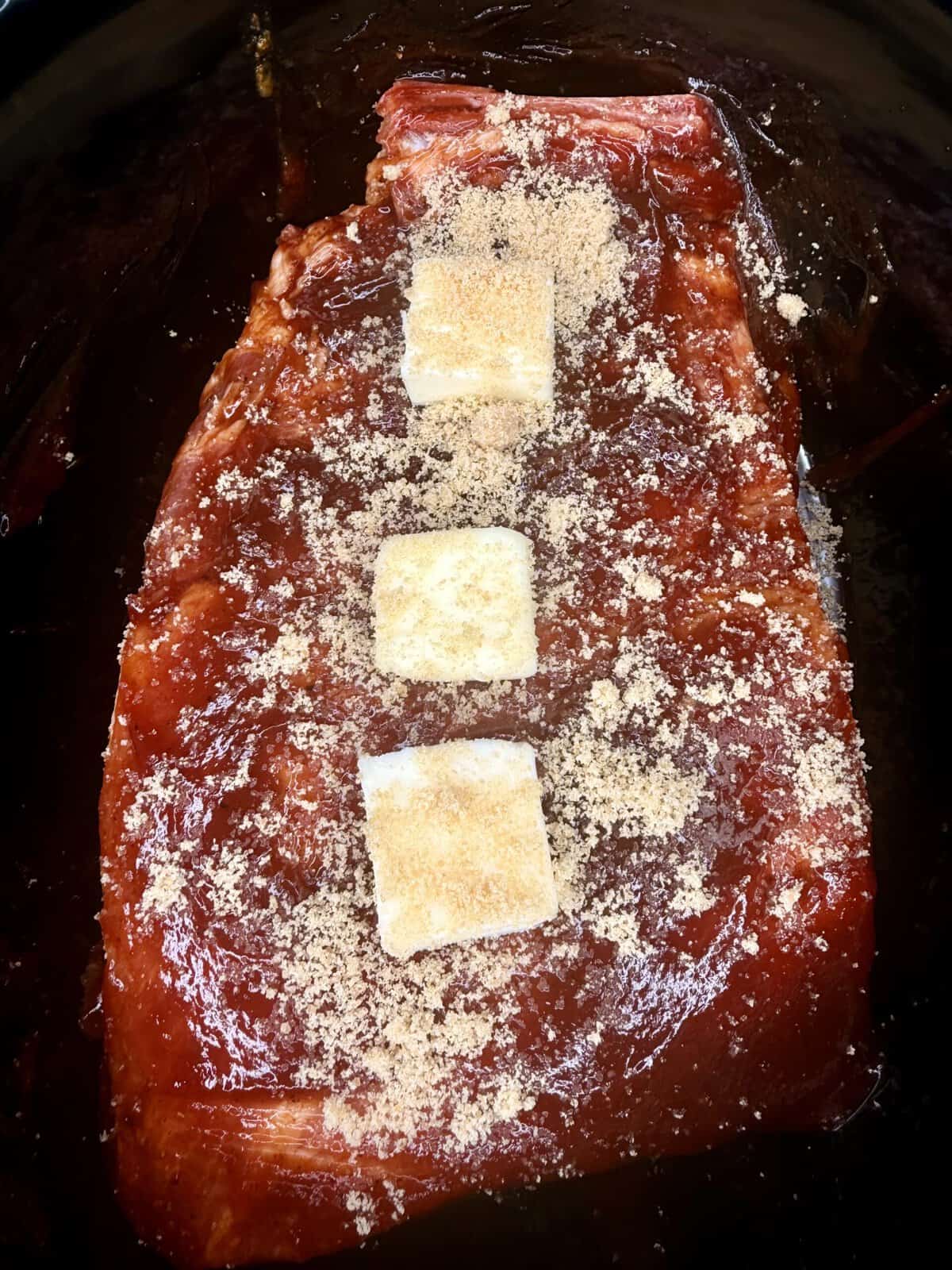 A slab of raw meat coated in a barbecue sauce, topped with three square pats of butter and a sprinkle of brown sugar, placed in a dark pan or slow cooker.