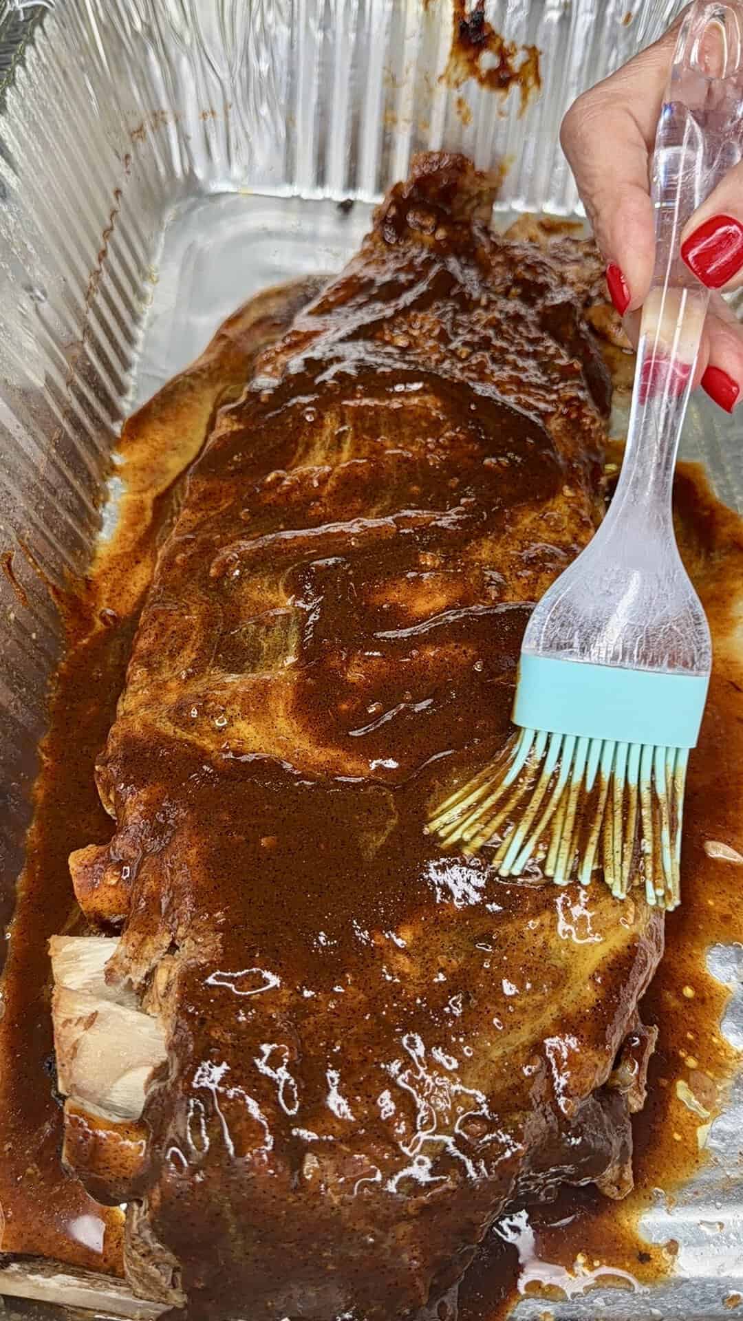 A hand with red nail polish uses a silicone brush to spread barbecue sauce over a rack of ribs in a foil tray.