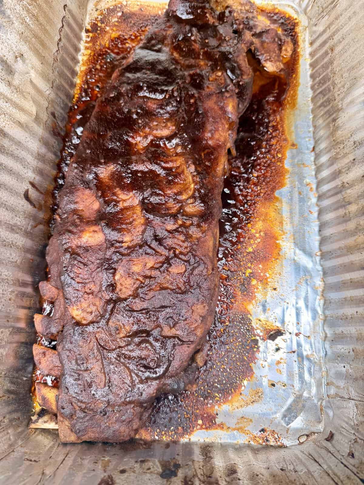 A rack of barbecued ribs coated in a dark, glossy sauce sits in a disposable aluminum tray with juices pooling around it. The ribs appear tender and well-cooked with a caramelized crust.