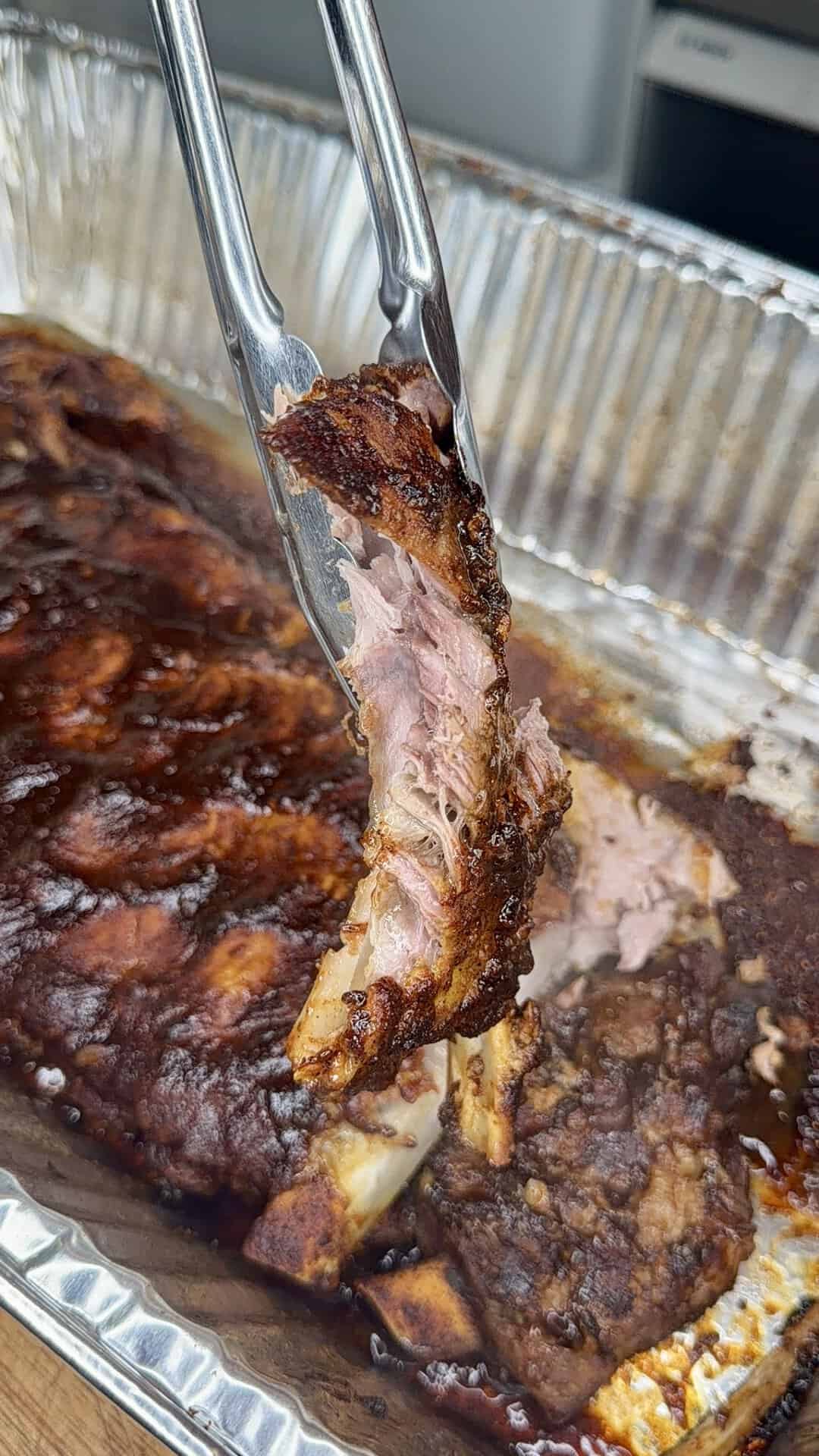 Close-up of metal tongs holding a tender, saucy piece of barbecued meat above a disposable foil tray filled with more barbecue ribs. The meat looks juicy and cooked, with a crispy, charred exterior.