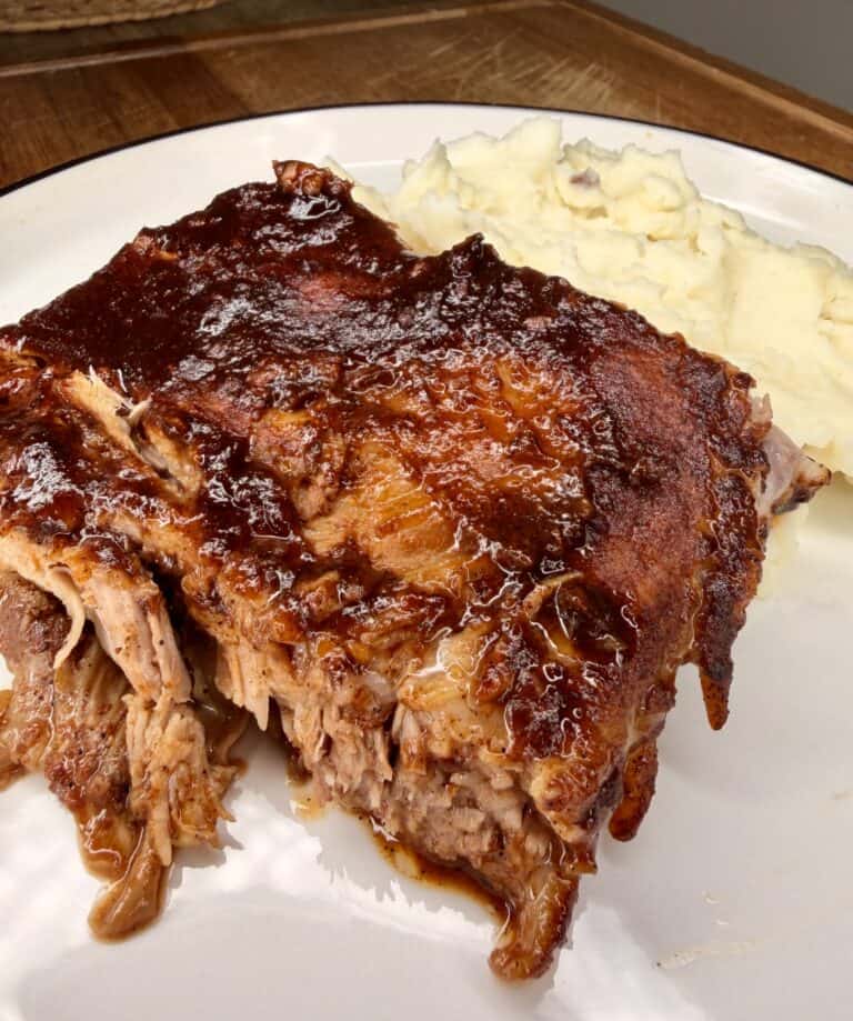 A plate of glazed, barbecued ribs with a side of creamy mashed potatoes on a white plate, set on a wooden table.
