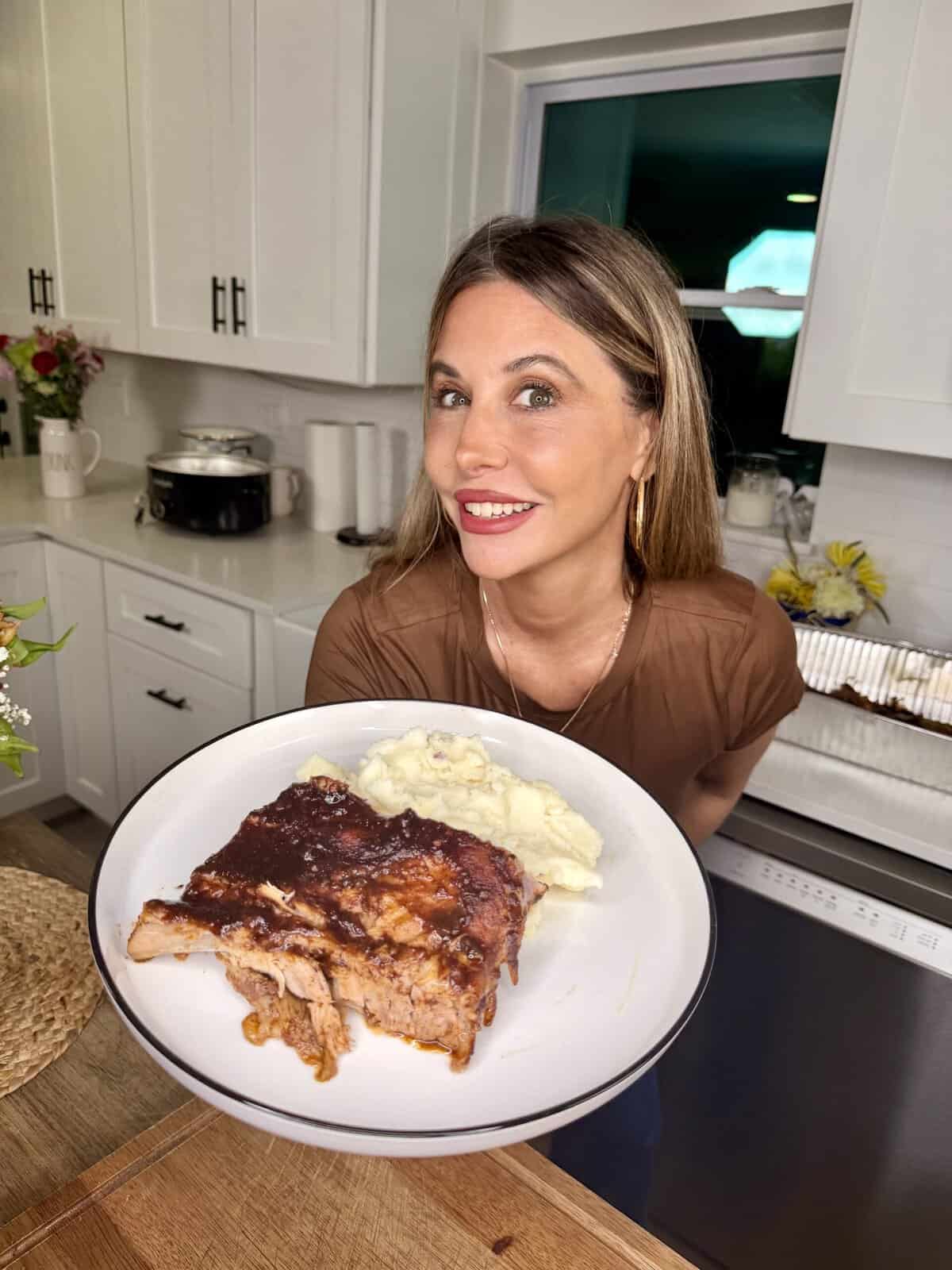 A woman in a kitchen smiles while holding a plate with barbecue ribs and mashed potatoes. White cabinets and flowers are visible in the background.