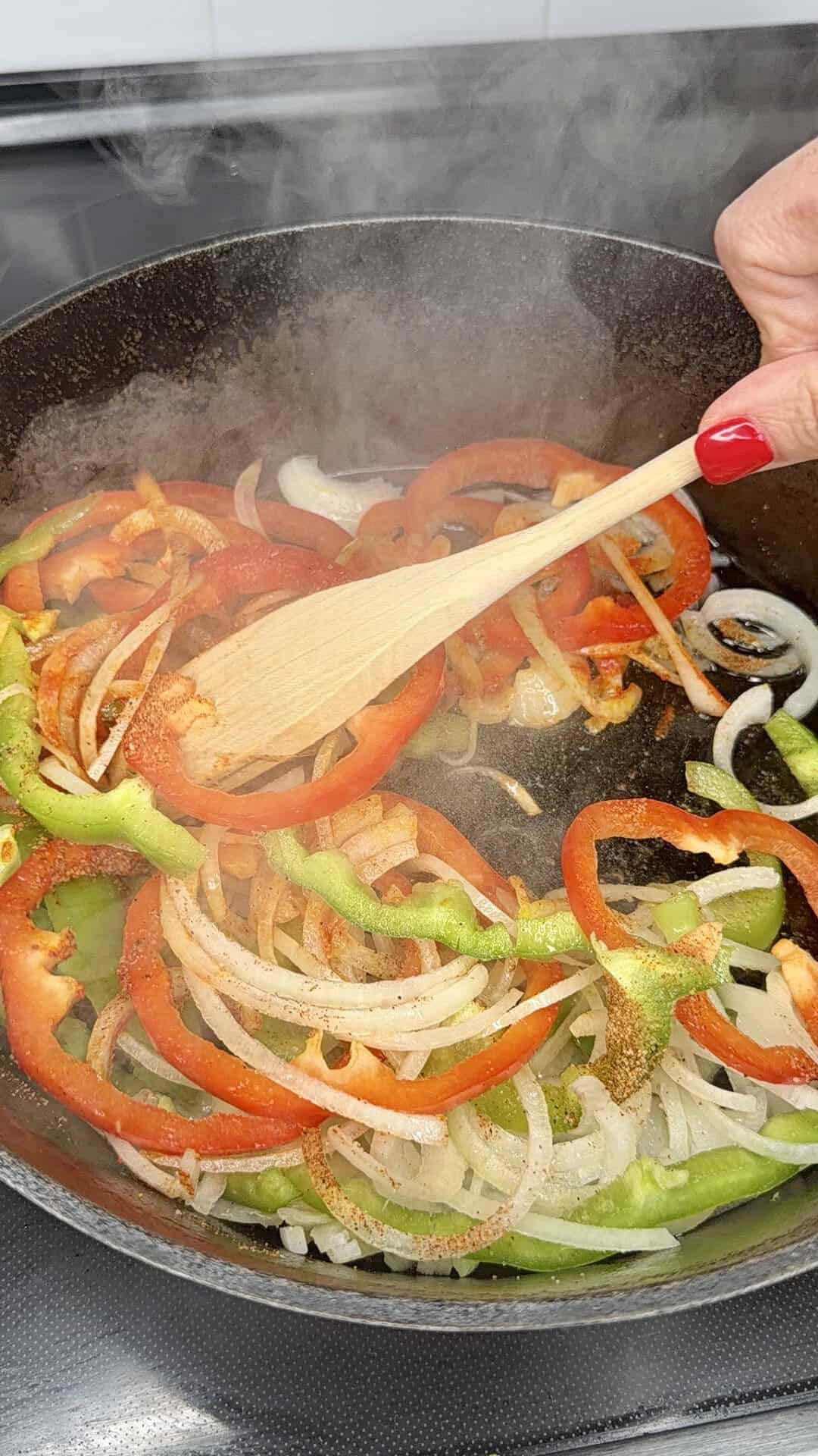 A hand stirs sliced red and green bell peppers and onions in a steaming pan with a wooden spatula.