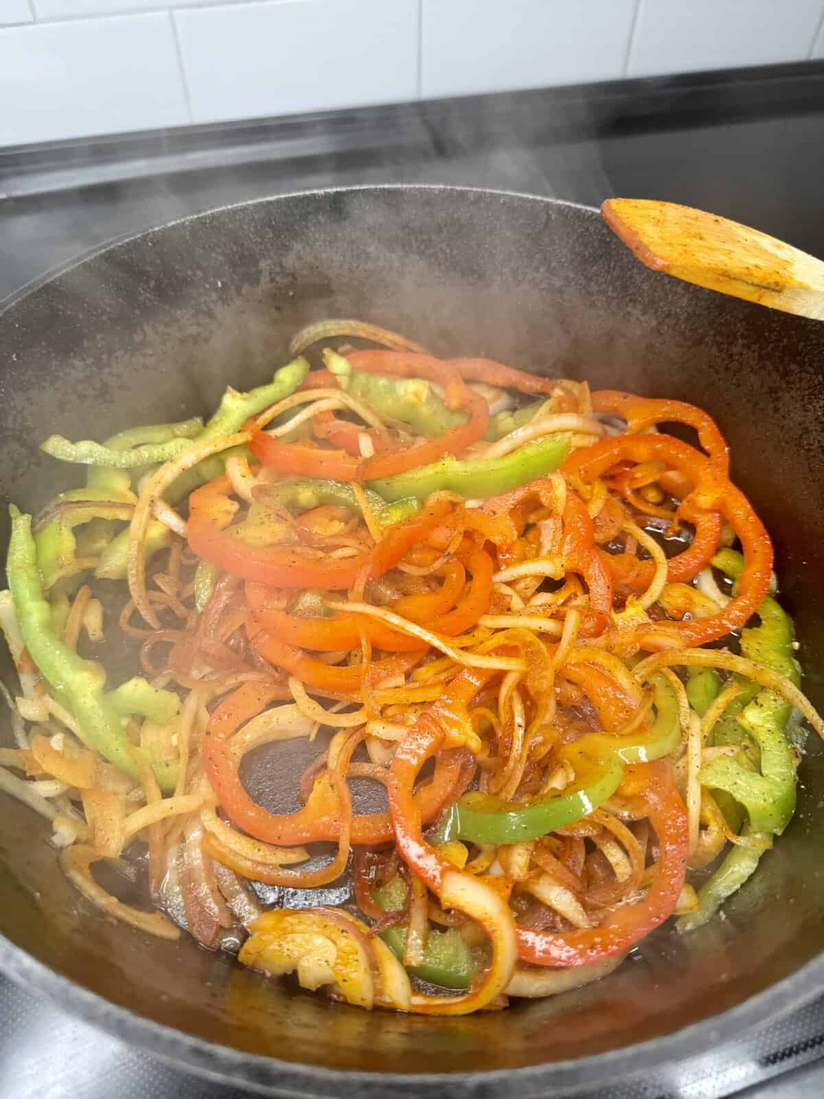 Colorful sliced red and green bell peppers and onions are being sautéed in a black cast iron skillet, with steam rising and a wooden spatula visible to the right.