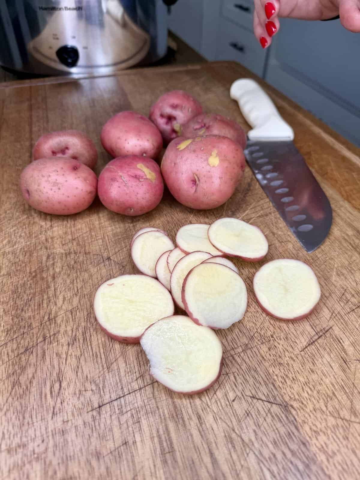 A hand holds a knife above a wooden cutting board with whole and sliced red potatoes. A slow cooker and kitchen counter are in the background.