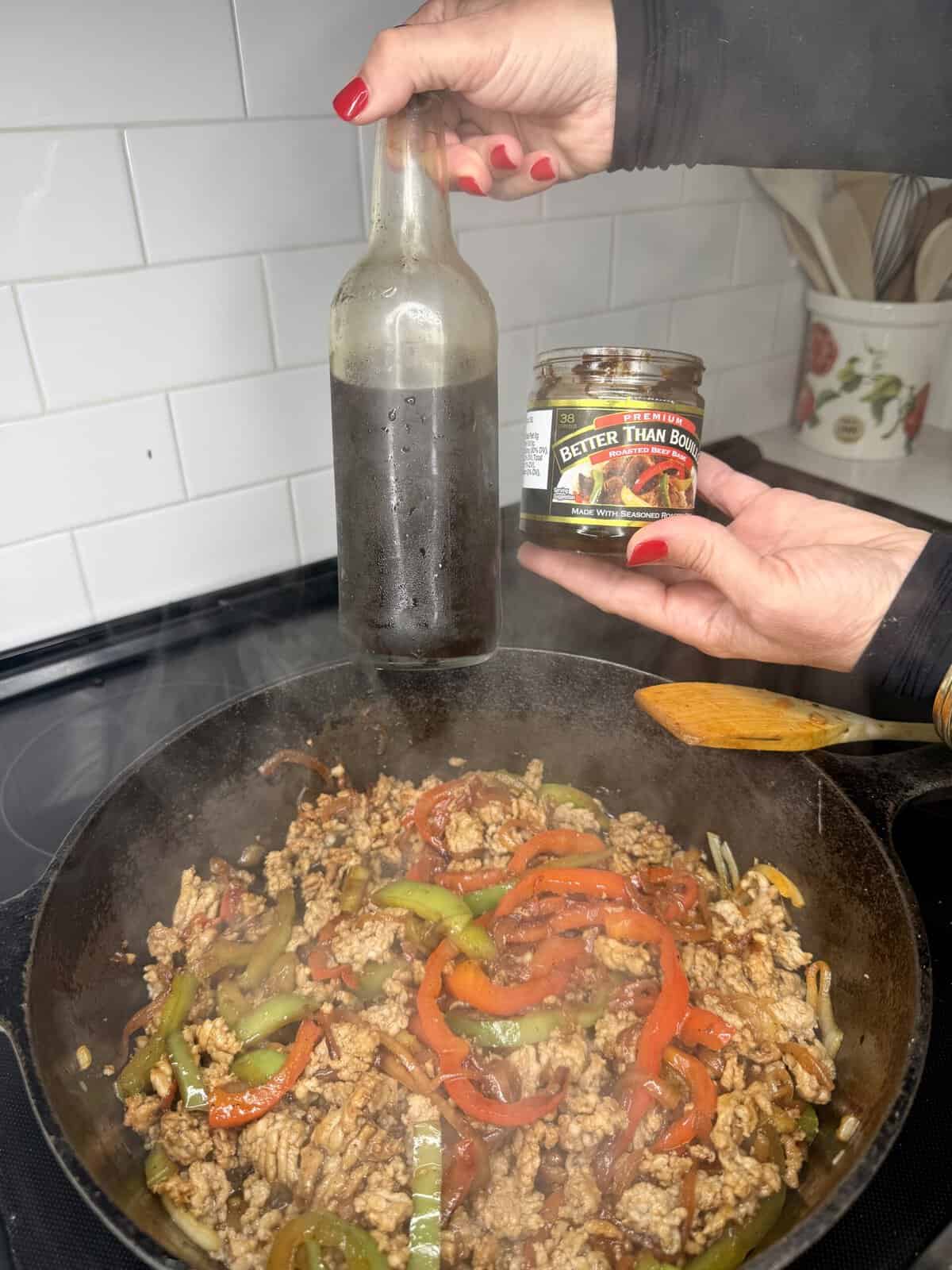 A person holds a bottle and a jar of Better Than Bouillon over a pan of cooked ground meat, onions, and bell peppers on a stovetop. Steam rises from the pan.