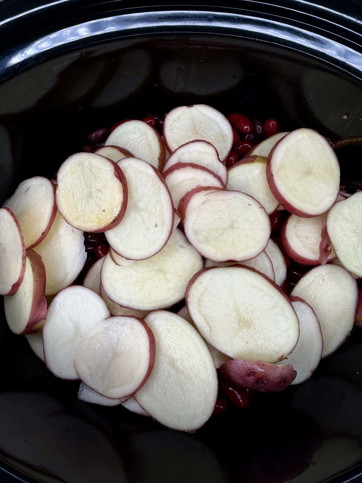 Sliced red potatoes layered on top of cranberries inside a black slow cooker, ready to be cooked.