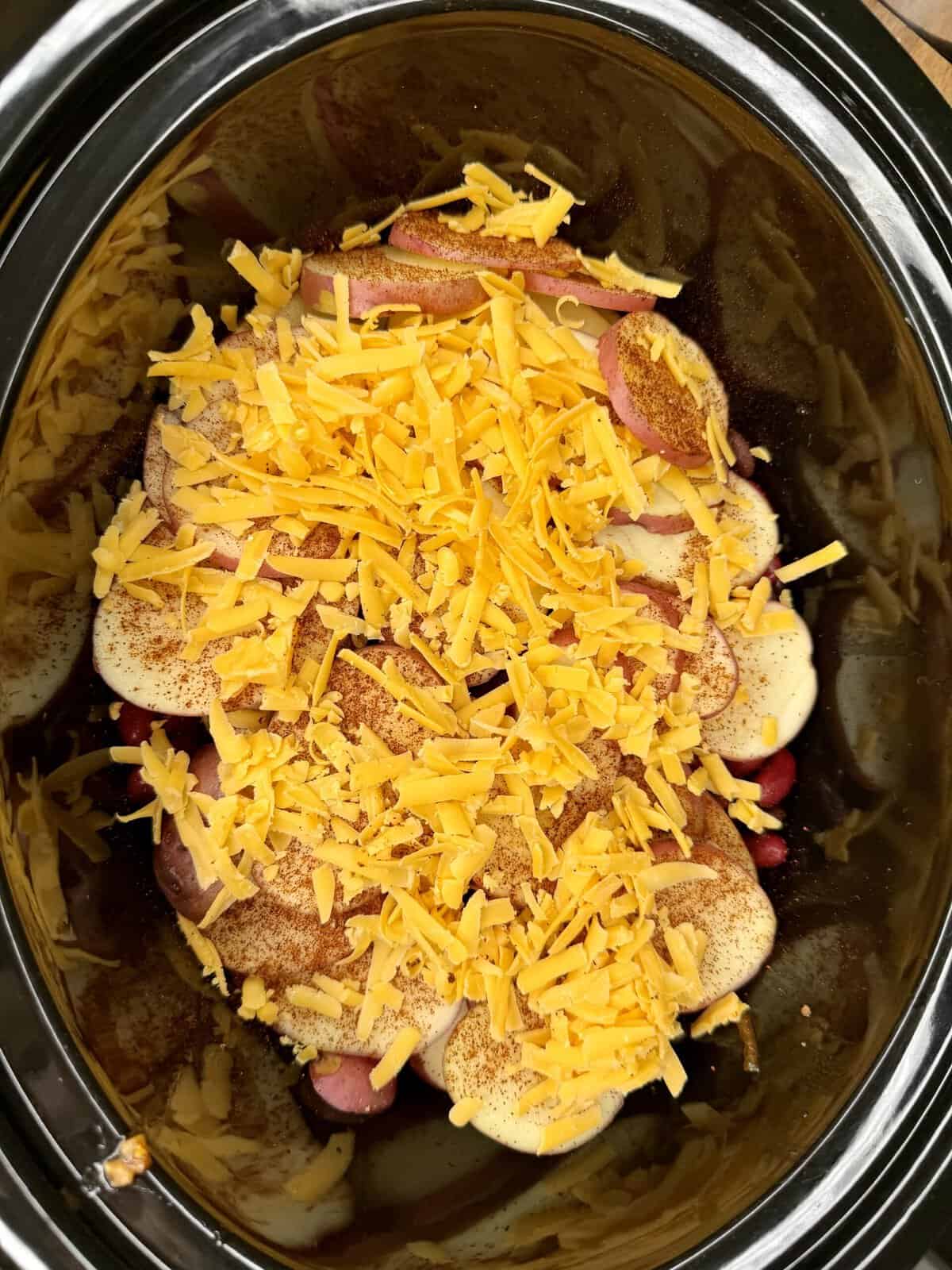 A slow cooker filled with sliced potatoes, sliced sausage, shredded cheddar cheese, and sprinkled seasoning, ready to be cooked.