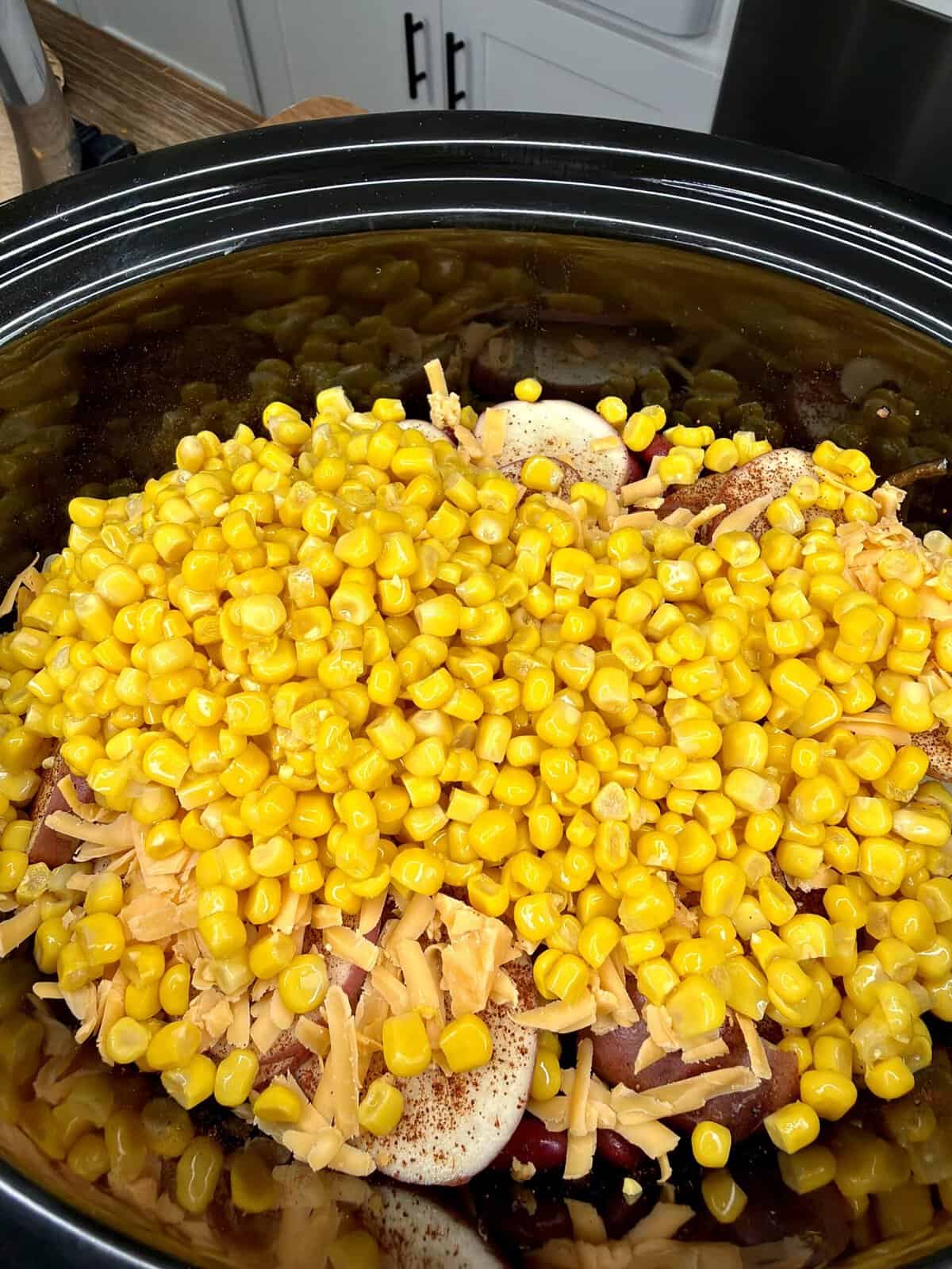 A slow cooker filled with sliced potatoes, shredded cheddar cheese, and a generous layer of yellow corn, ready to be cooked.