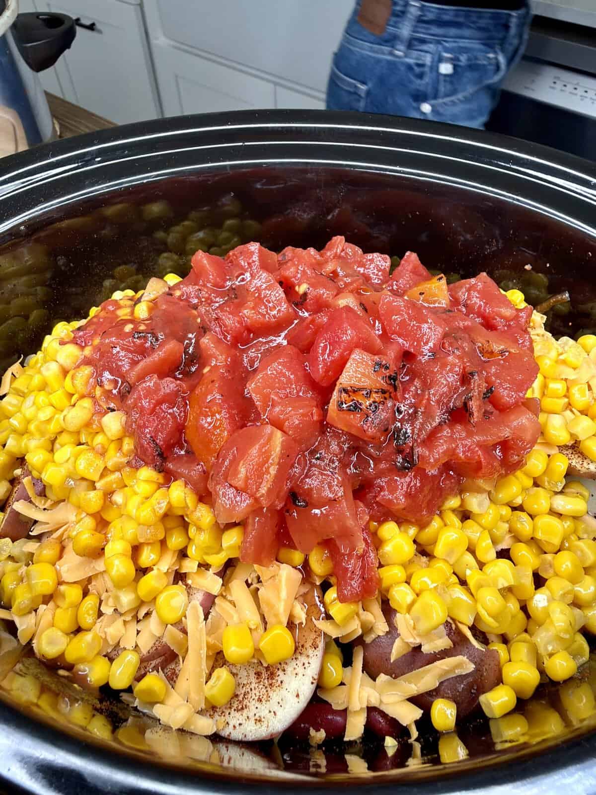 A slow cooker filled with layers of corn, shredded cheddar cheese, and diced tomatoes. In the background, a person in jeans stands near a kitchen counter.