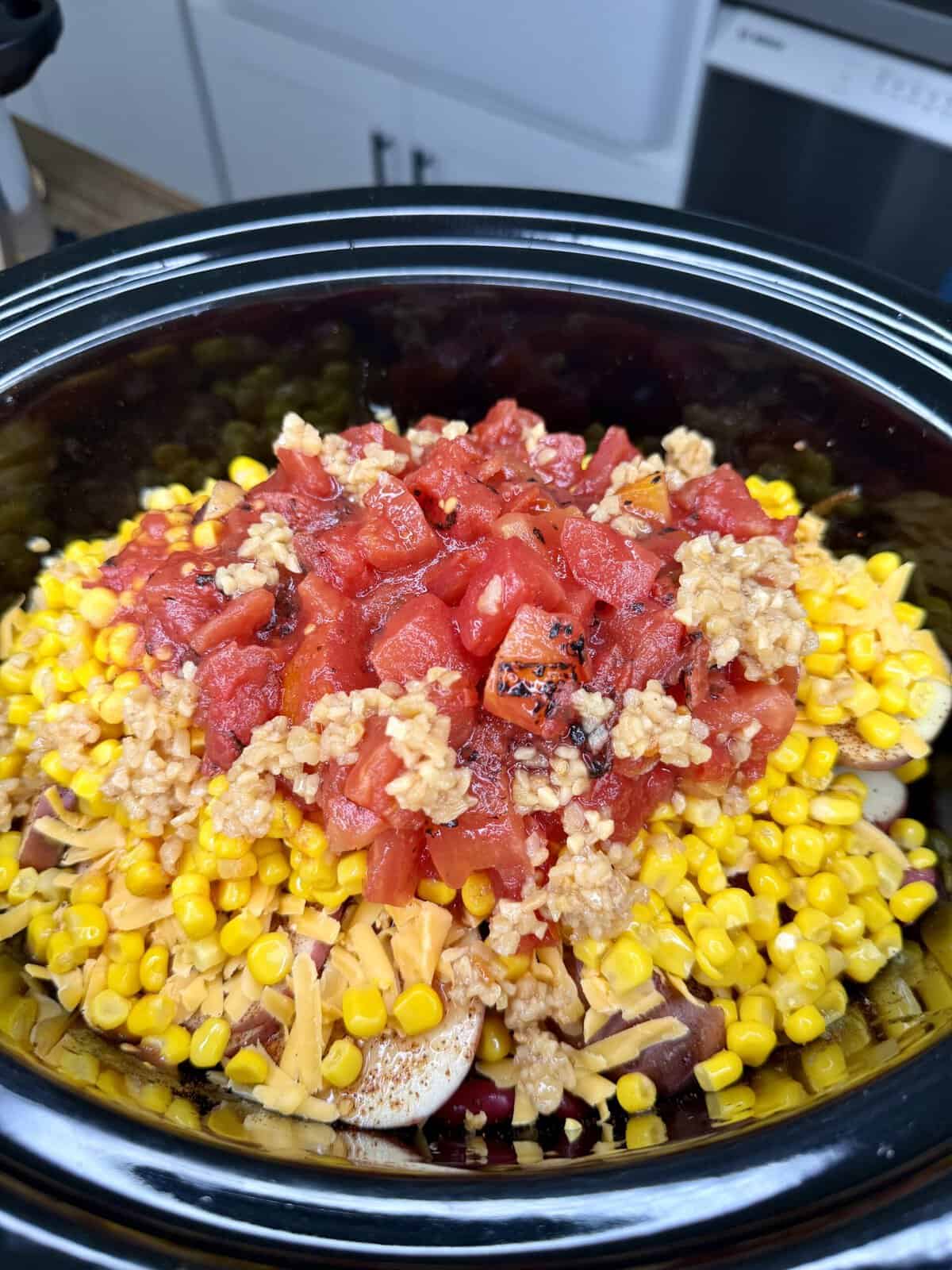 A close-up of a slow cooker filled with corn, diced tomatoes, minced garlic, and other ingredients, ready to be cooked. Kitchen counters and cabinets are visible in the background.