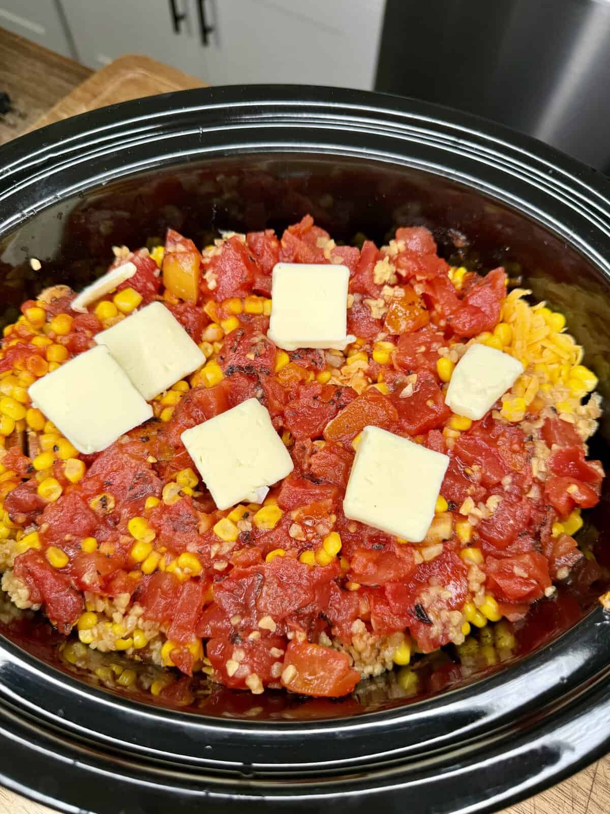 A slow cooker filled with a mixture of corn, tomatoes, pasta, and ground meat, topped with five square pats of butter, ready to be cooked.