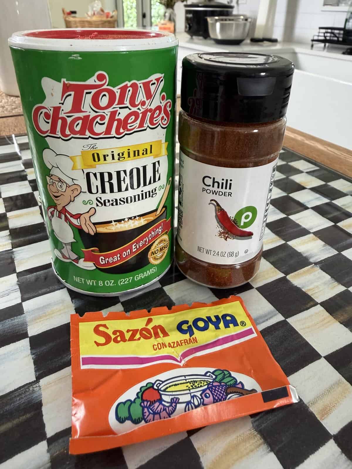 A can of Tony Chachere’s Creole Seasoning, a jar of Publix Chili Powder, and a packet of Sazón Goya seasoning sit on a checkered table. A kitchen counter with various items is visible in the background.