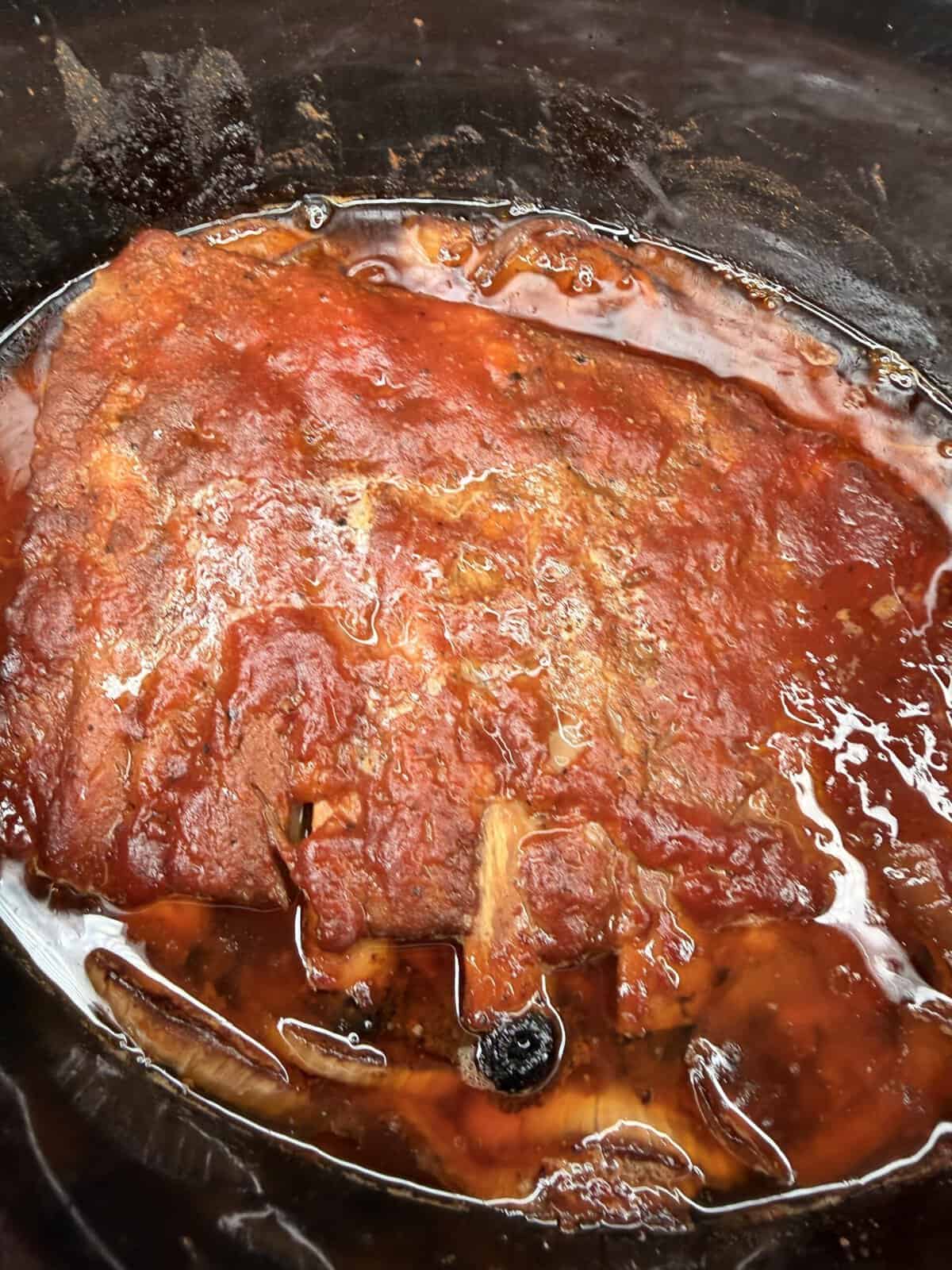 A close-up of a slab of meat covered in a red tomato-based sauce, cooking in a slow cooker or crockpot with visible juices and seasonings.