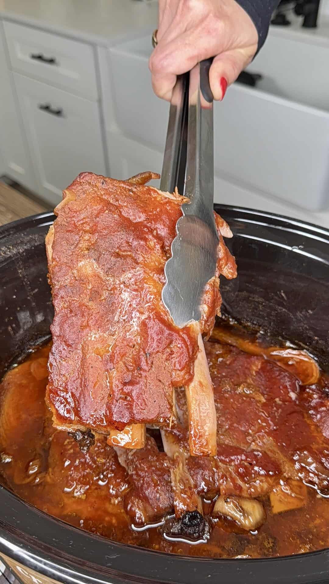 A hand holding metal tongs lifts a saucy, cooked rack of ribs from a slow cooker filled with barbecue sauce in a bright kitchen.