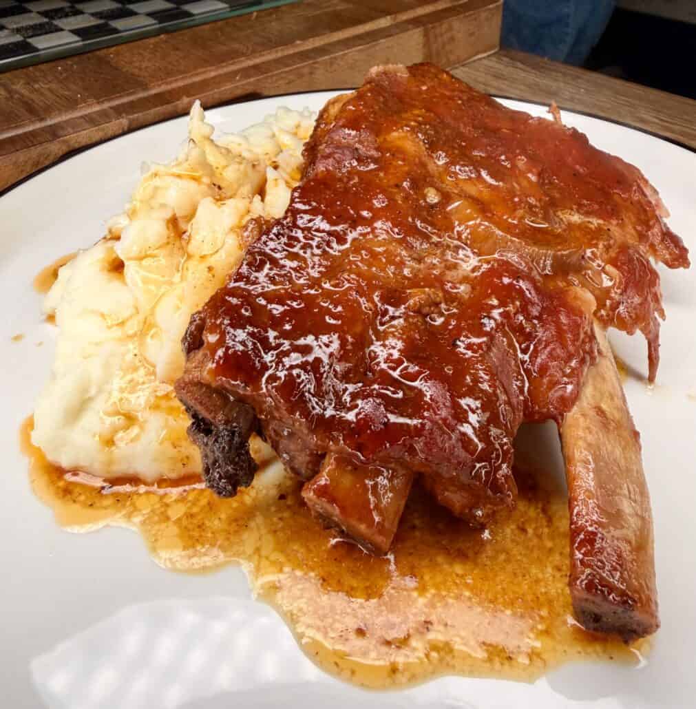 A plate of saucy barbecue ribs served on top of creamy mashed potatoes, with some sauce drizzled around the food. The dish is presented on a white plate with a dark rim.