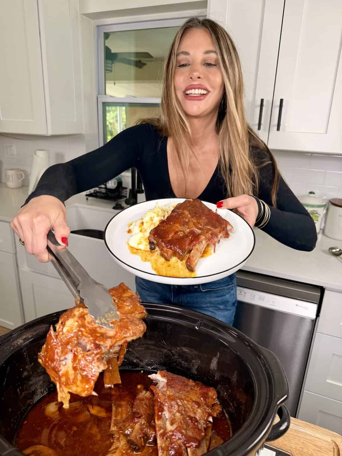 A smiling woman in a black shirt serves saucy ribs from a slow cooker onto a plate of mashed potatoes in a bright kitchen.