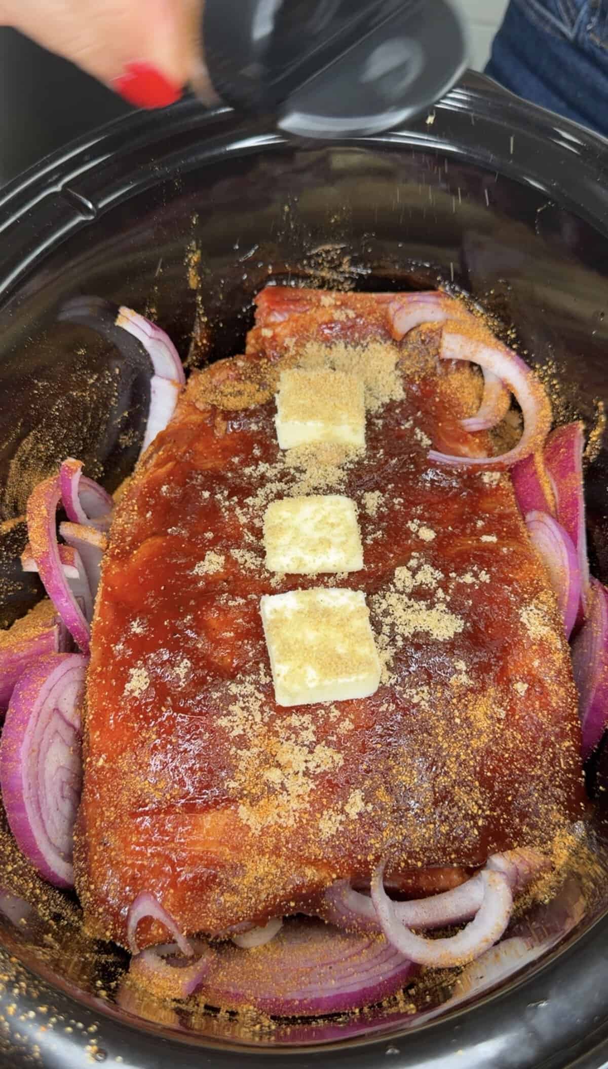 A slab of seasoned meat topped with three pats of butter and brown sugar sits on a bed of sliced red onions in a slow cooker, ready to be cooked.