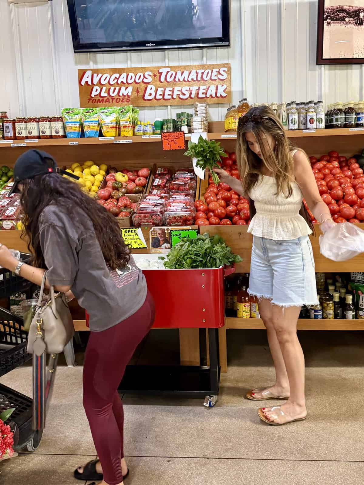 Two women shopping for produce at a grocery store stand with tomatoes, avocados, and other fruits. One woman bends over her cart, while the other inspects leafy greens. Signs label various produce items above them.