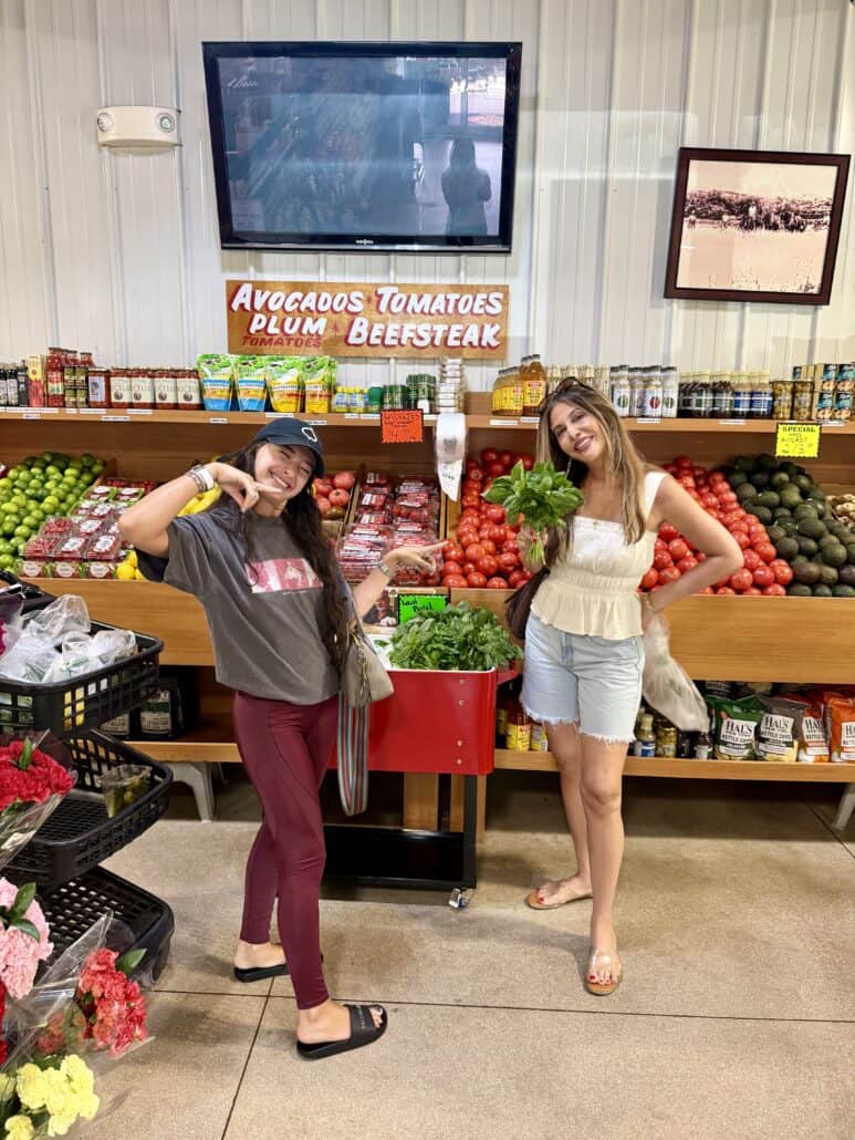 Two women smile and pose in front of a colorful fruit and vegetable display at a market. One holds greens, the other gestures with her hand. Signs for avocados, tomatoes, and other produce are visible.