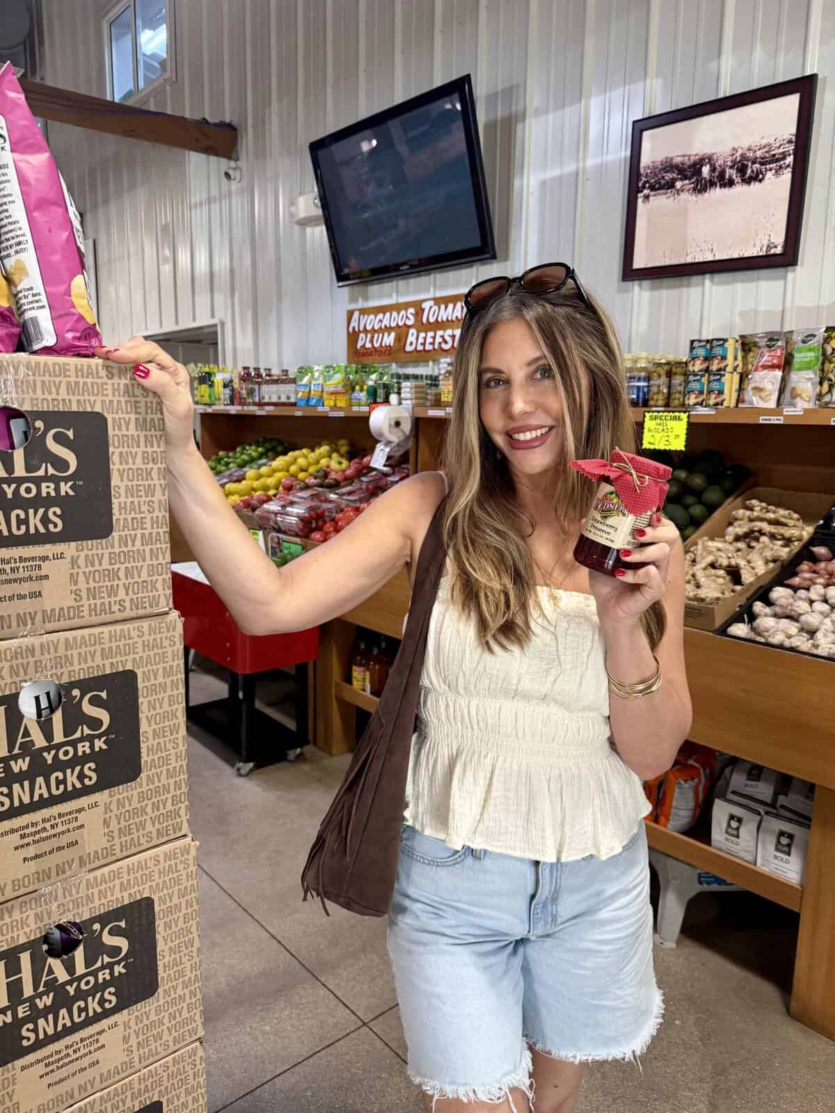 A woman in a white top and denim shorts holds a jar while shopping in a grocery store filled with fresh produce, snacks, and other items. She is smiling and standing next to a display of chips.