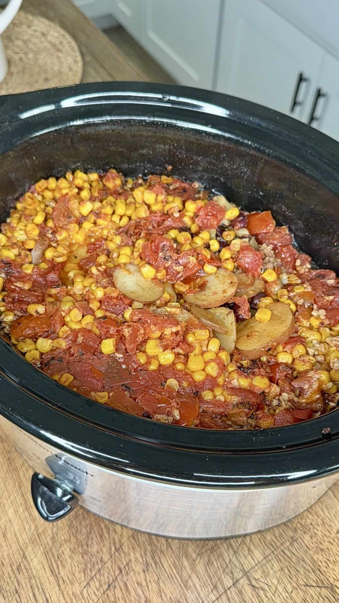 A slow cooker filled with a stew containing corn, tomatoes, sliced potatoes, and ground meat, sitting on a wooden countertop in a kitchen.