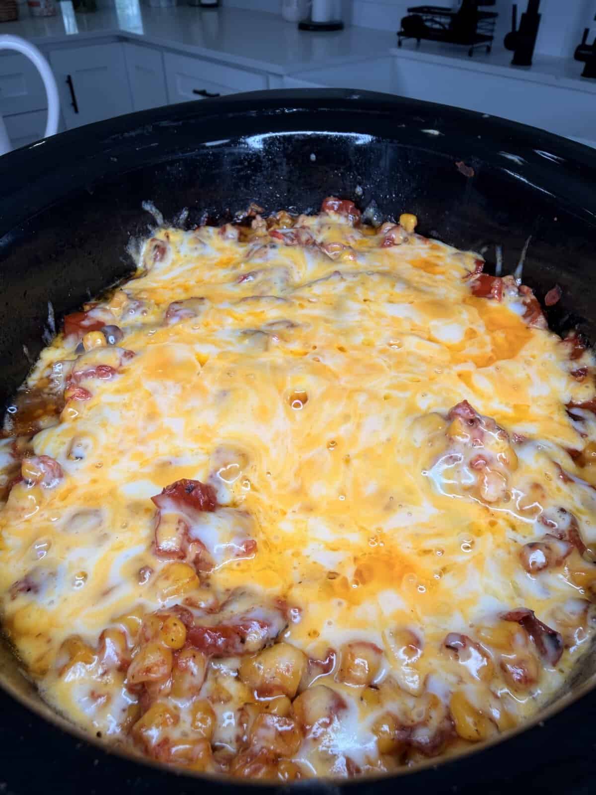 A slow cooker filled with a cheesy, melted casserole featuring mixed ingredients, likely beans and meat, in a kitchen setting.