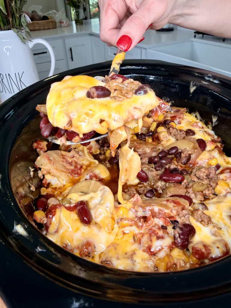 A hand with red nails lifts a cheesy serving of taco casserole from a slow cooker, showing beans, corn, ground meat, and melted cheese in a kitchen setting.