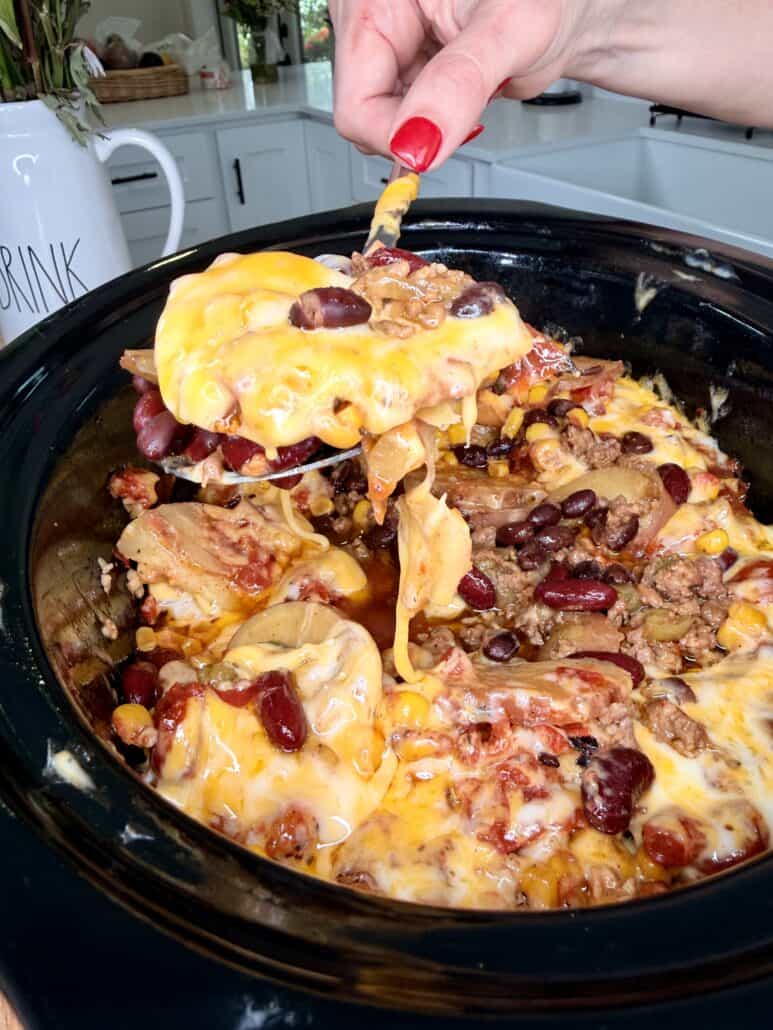 A hand with red nails lifts a cheesy serving of taco casserole from a slow cooker, showing beans, corn, ground meat, and melted cheese in a kitchen setting.