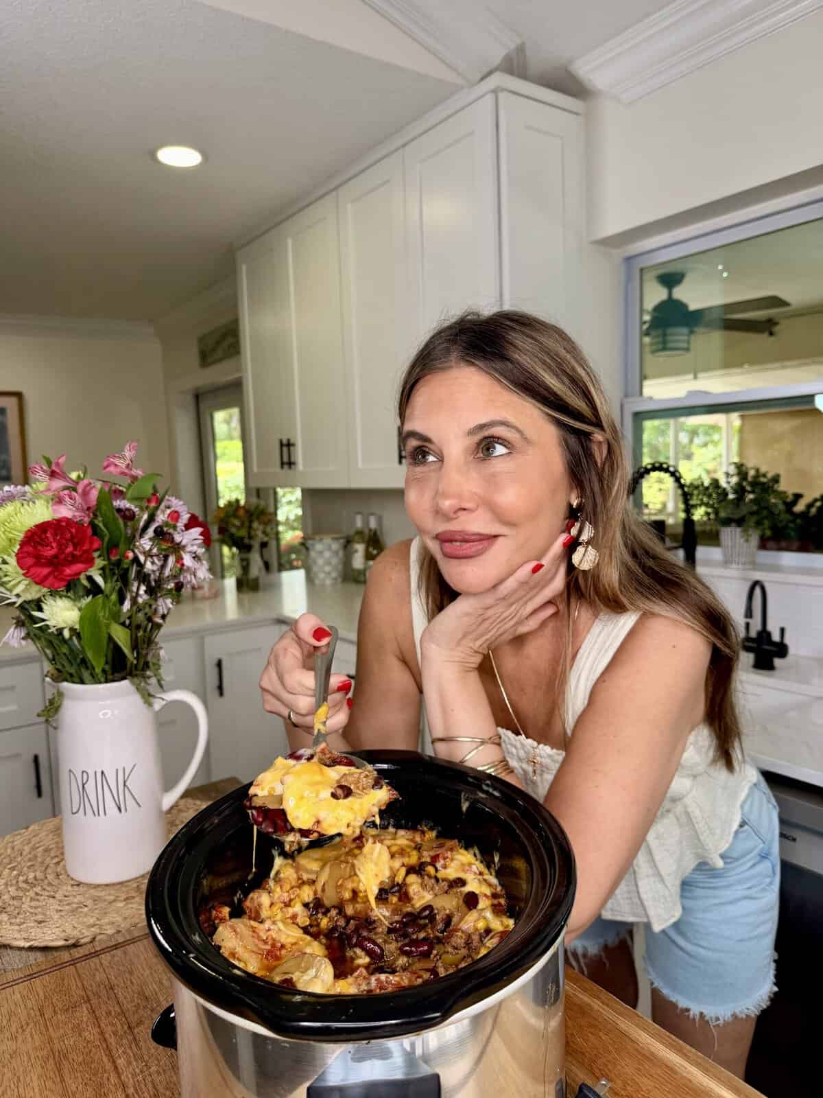 A woman in a kitchen smiles while holding a spoonful of cheesy casserole from a slow cooker. There is a vase of flowers labeled DRINK on the counter beside her.