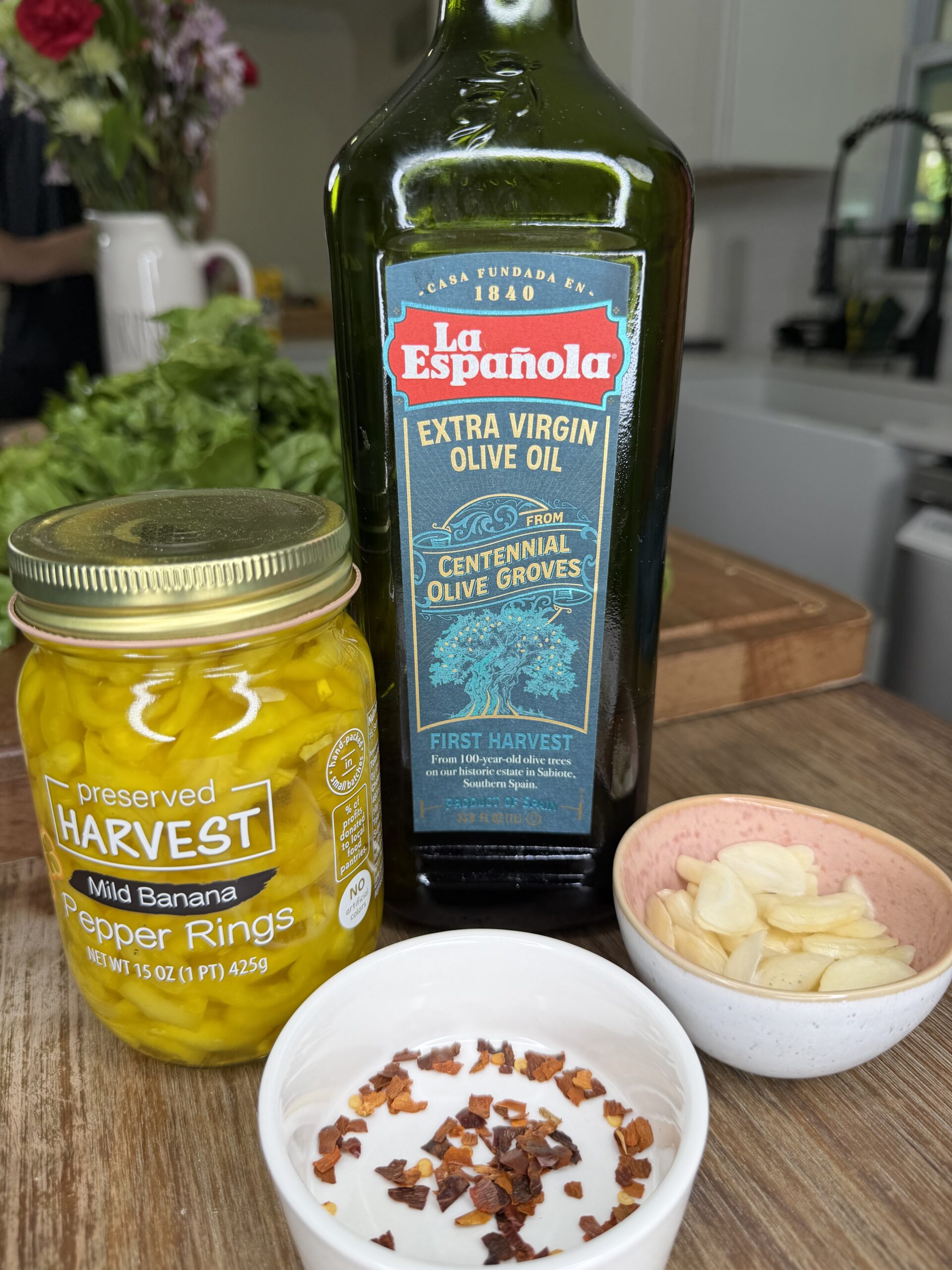 A jar of mild banana pepper rings, a bottle of La Española extra virgin olive oil, a bowl of sliced garlic, a bowl of red pepper flakes, and greens on a kitchen counter.