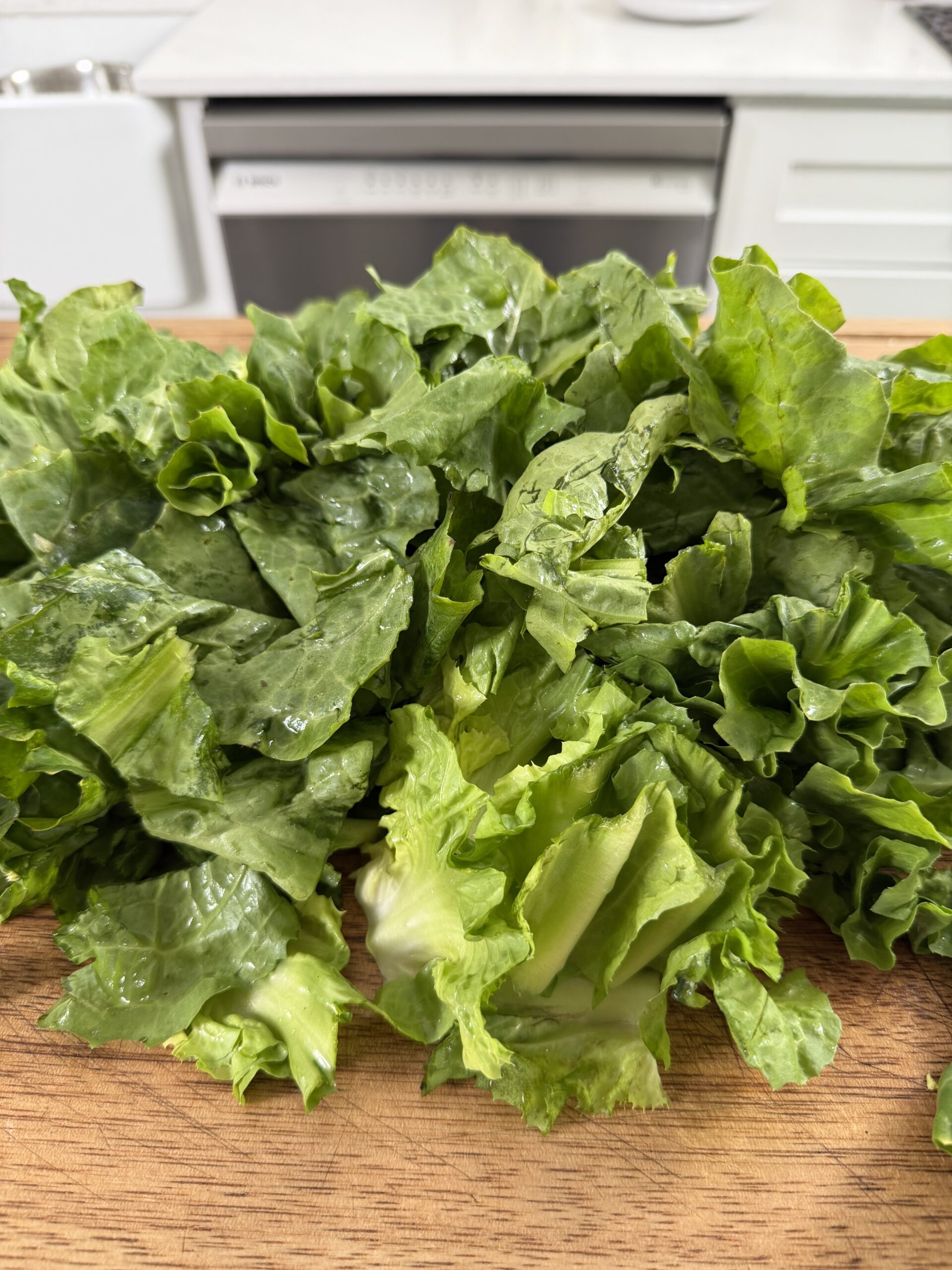 A pile of fresh, chopped green lettuce sits on a wooden cutting board in a kitchen with white cabinets and a dishwasher in the background.