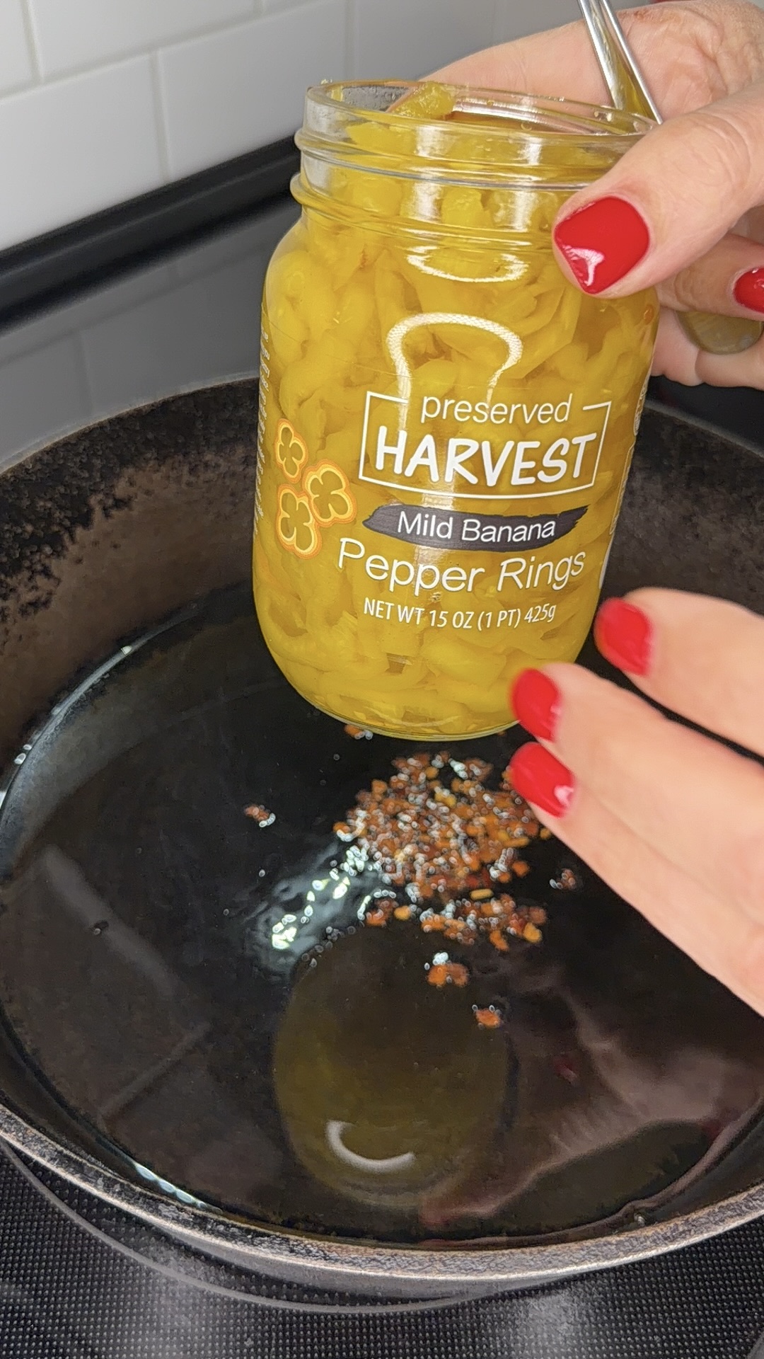 A hand holds a jar of mild banana pepper rings above a skillet with oil and red pepper flakes. The person has red nail polish and is about to add the peppers to the pan.