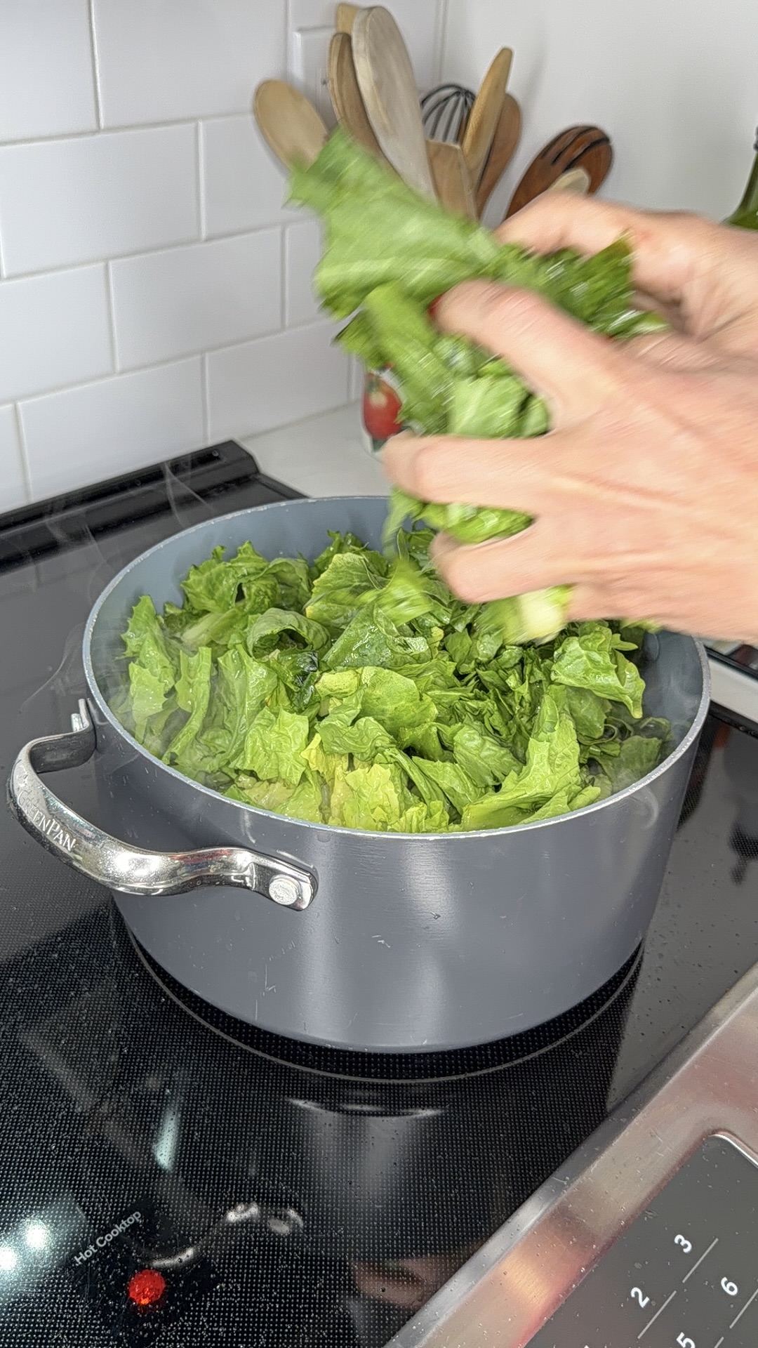 A person adds fresh green leafy vegetables to a pot on a stovetop, with wooden utensils and kitchen tiles visible in the background.