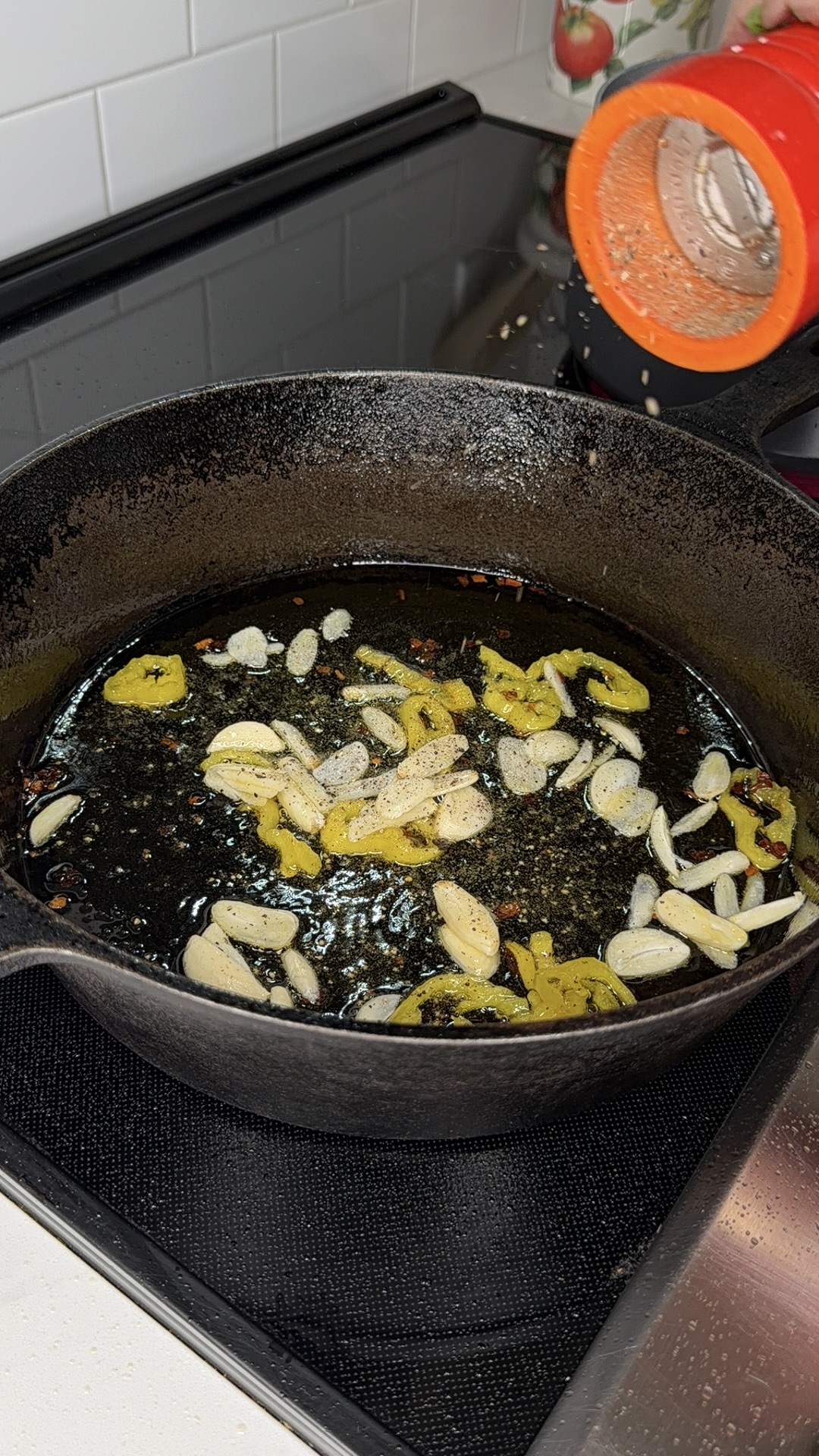 Sliced garlic and yellow peppers sizzling in oil in a black cast iron skillet on a stovetop. An orange spice container lies tipped over on the countertop nearby.