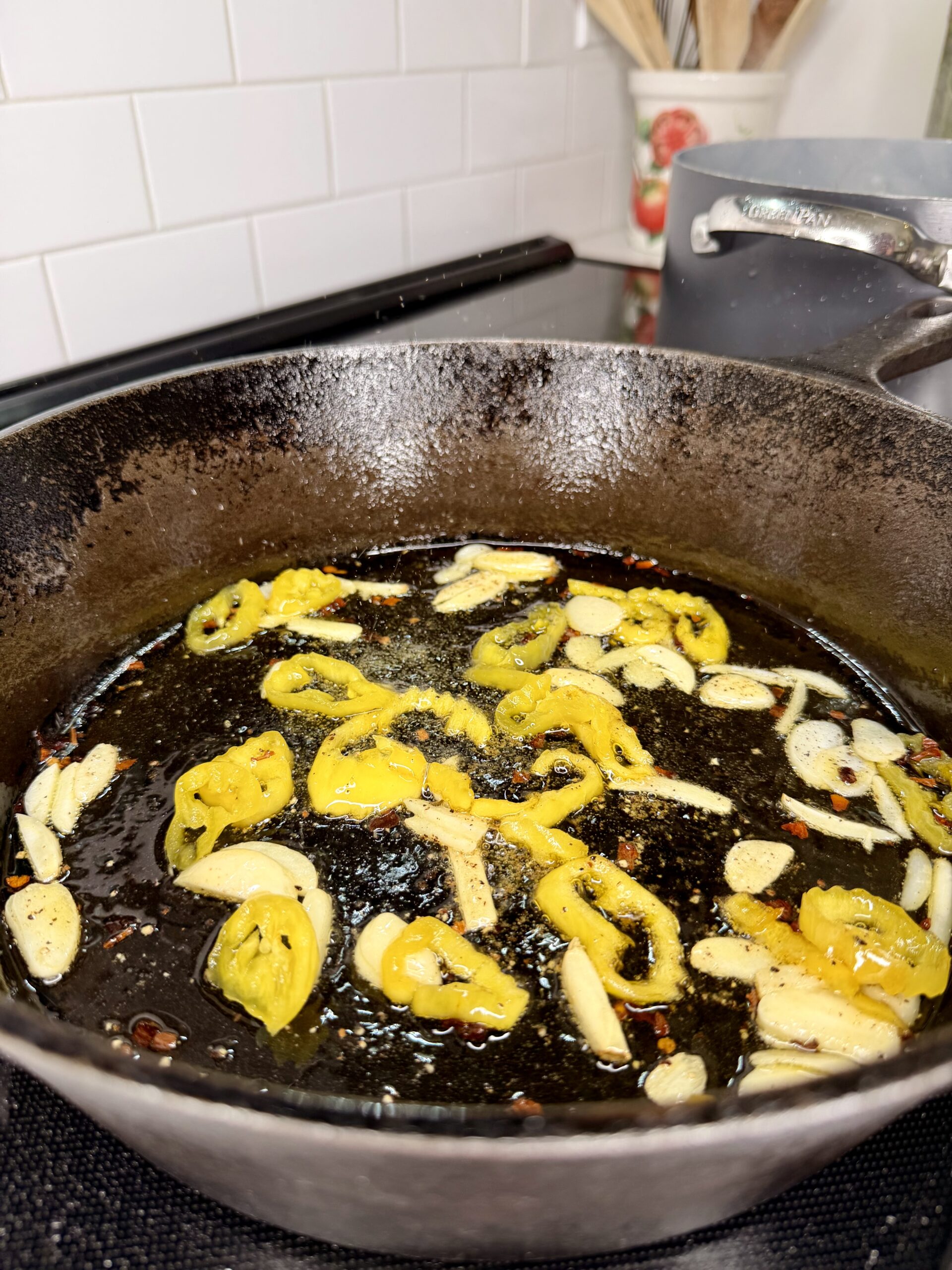 Sliced yellow peppers and garlic are sizzling in oil in a black cast iron skillet on a stovetop, with a white tile backsplash and kitchen utensils in the background.