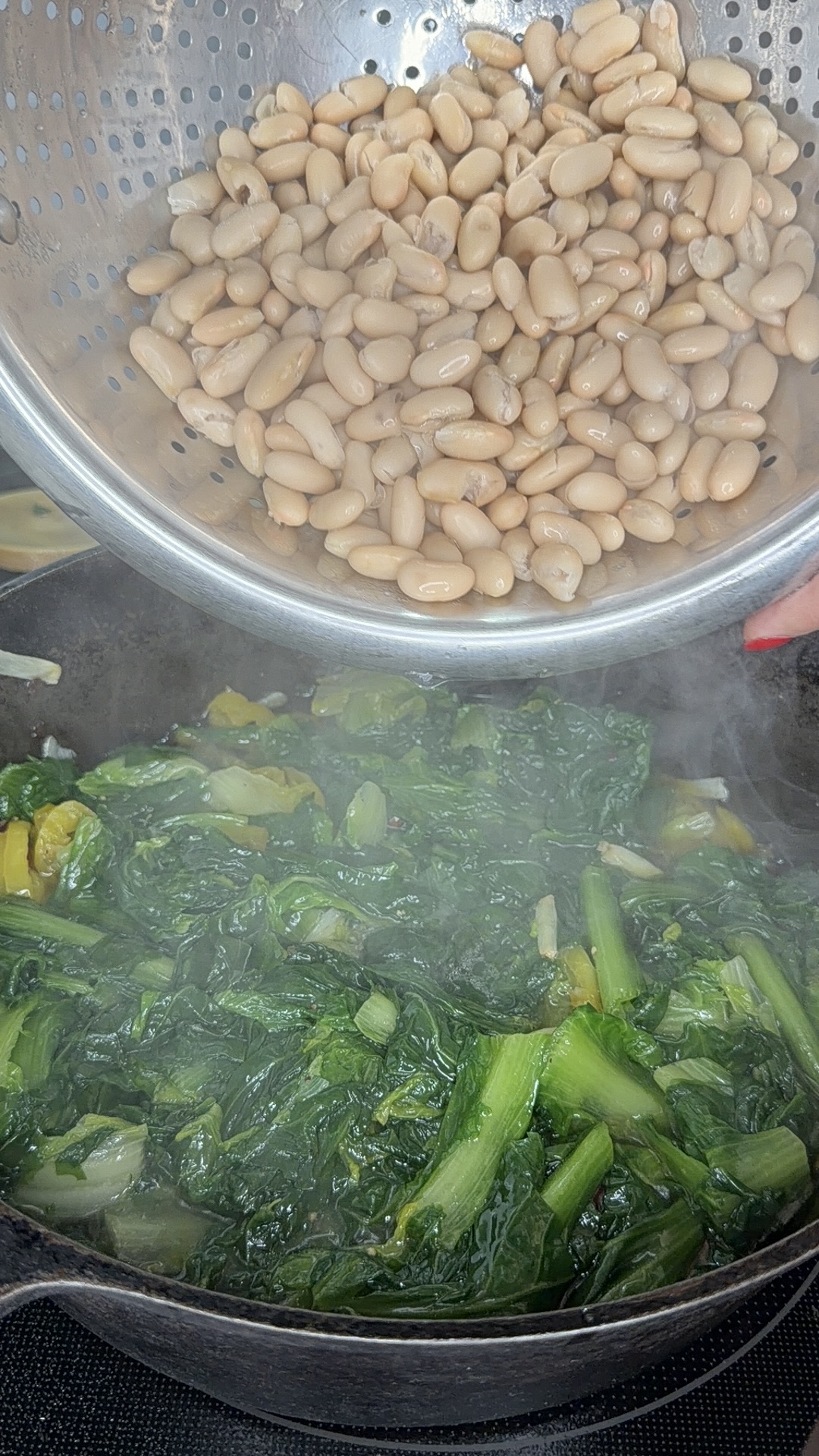 A colander of white beans is being poured into a cast iron skillet filled with cooked greens and vegetables, with steam rising from the pan.