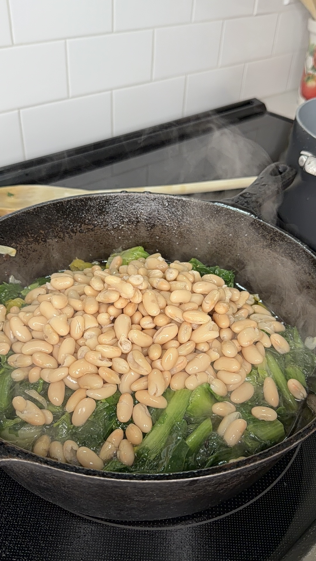 White beans and leafy greens cooking in a black cast iron skillet on a stovetop, with steam rising and a wooden spoon and pot visible in the background.