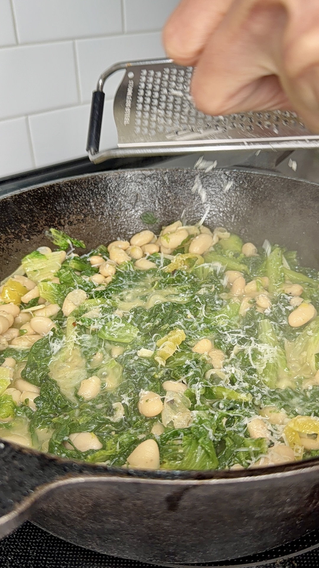 A hand grates cheese over a skillet filled with cooked white beans and leafy greens, likely kale, on a stovetop with a white tile backsplash.