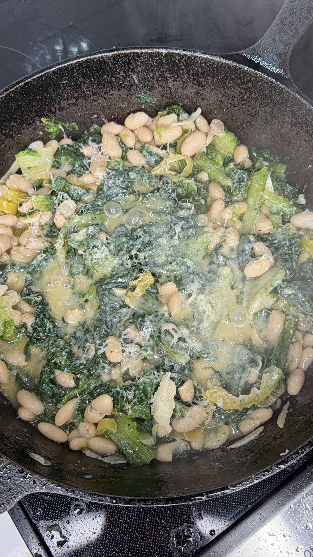 A cast iron skillet on a stove filled with white beans, wilted greens, and a light broth, cooking together.