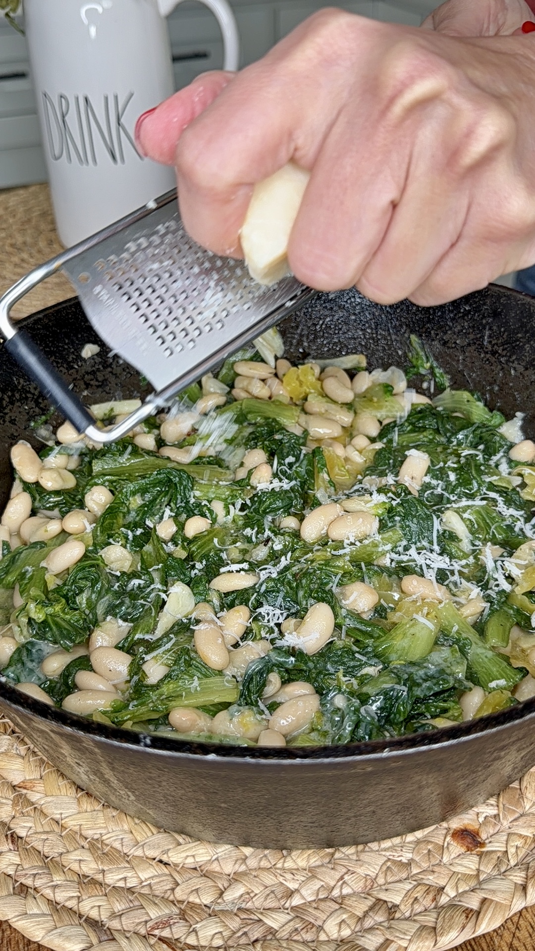 A hand grates cheese over a cast iron skillet filled with cooked greens and white beans. A white “DRINK” mug is visible in the background.