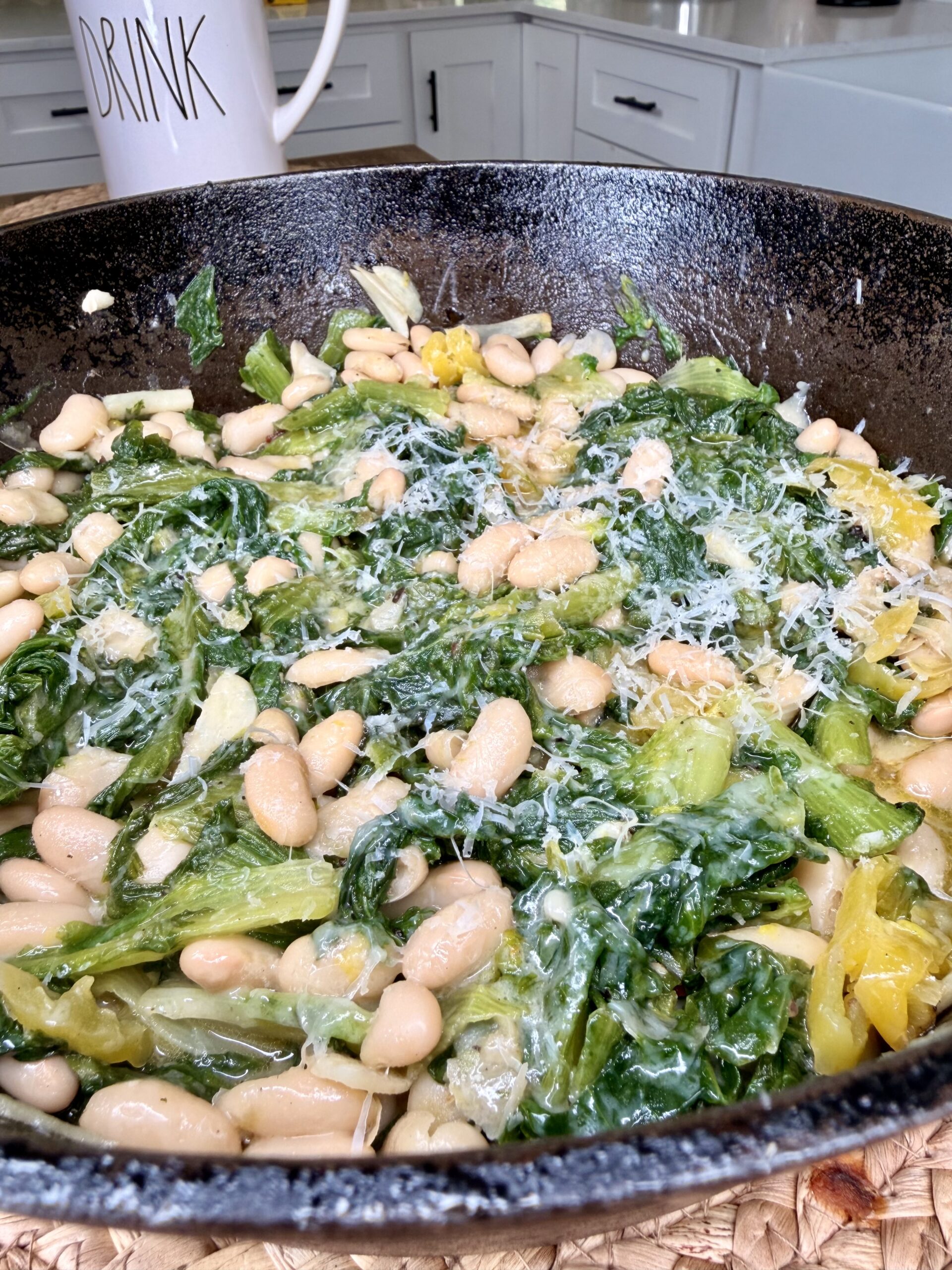 White beans, wilted greens, and grated cheese are cooked together in a black cast iron skillet. In the background, a white mug labeled “DRINK” sits on a kitchen counter.