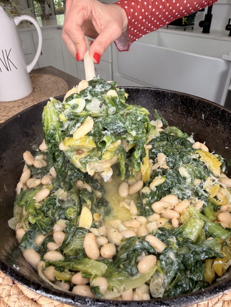 A hand in a red polka dot sleeve holds a spoonful of sautéed greens and white beans from a cast iron skillet in a bright kitchen.